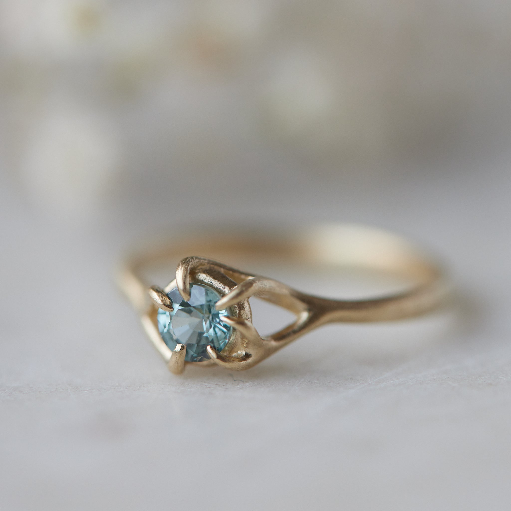 a round crystal blue montana sapphire is wrapped in gold branches. photographed on a white marble slab.
