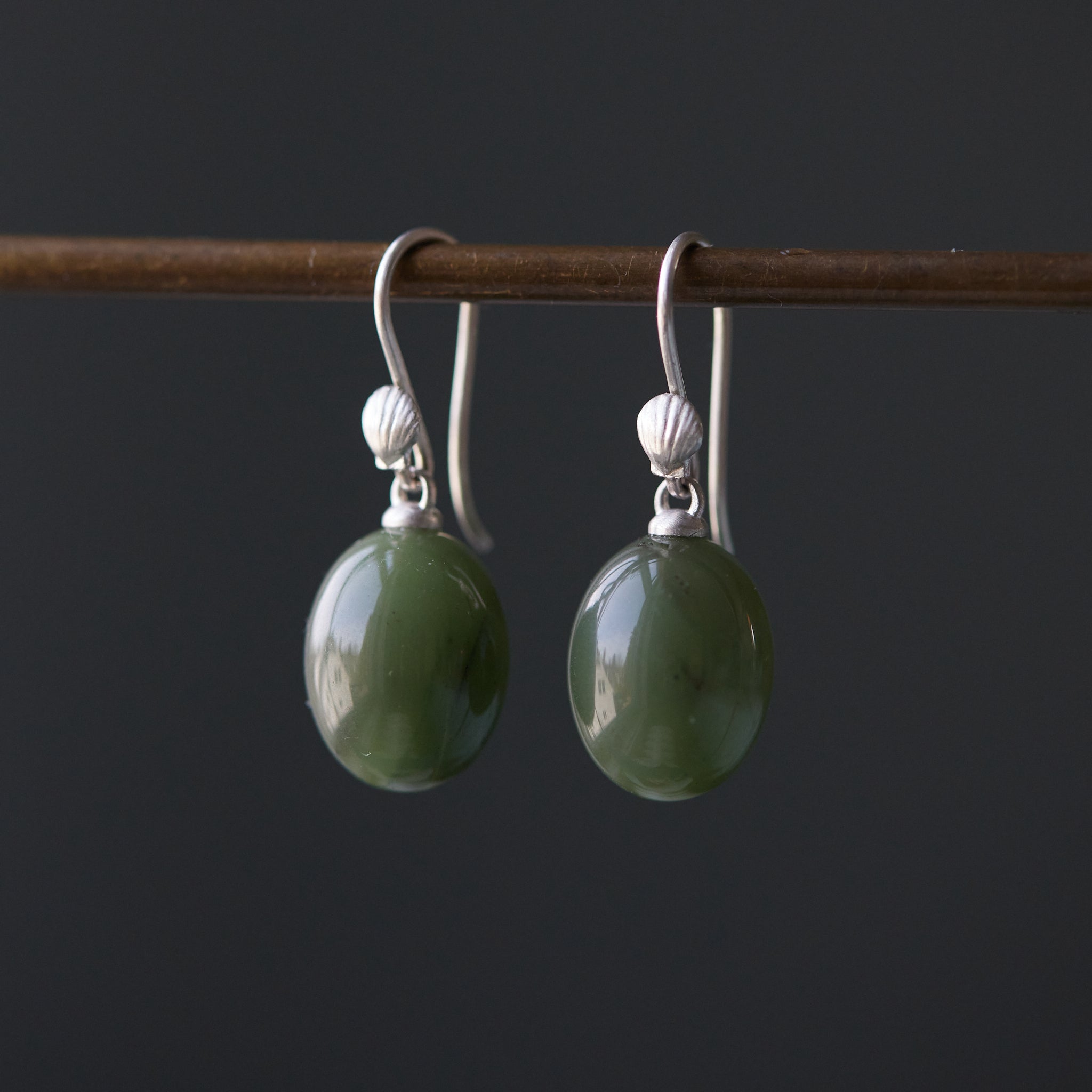 A set of earrings featuring silver or gold scallop shells attached to green jade spheres, displayed on a dark background.