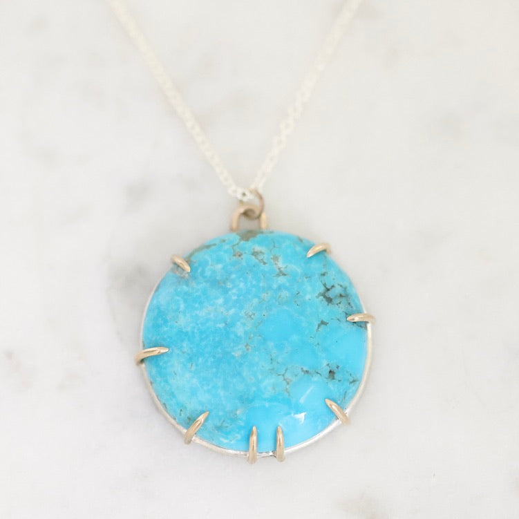 A round turquoise pendant necklace with a silver bezel and prong setting, resting on a white marble surface.