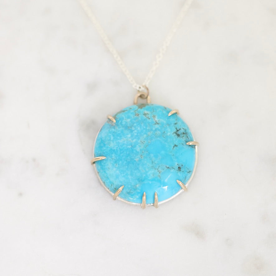 A round turquoise pendant necklace with a silver bezel and prong setting, resting on a white marble surface.
