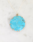 A round turquoise pendant necklace with a silver bezel and prong setting, resting on a white marble surface.