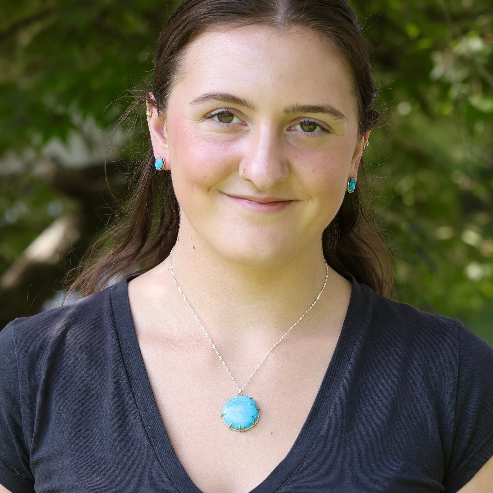 A woman with brown hair wearing a silver necklace with a round turquoise pendant and matching turquoise stud earrings, photographed outdoors against a blurred green foliage background.
