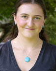 A woman with brown hair wearing a silver necklace with a round turquoise pendant and matching turquoise stud earrings, photographed outdoors against a blurred green foliage background.