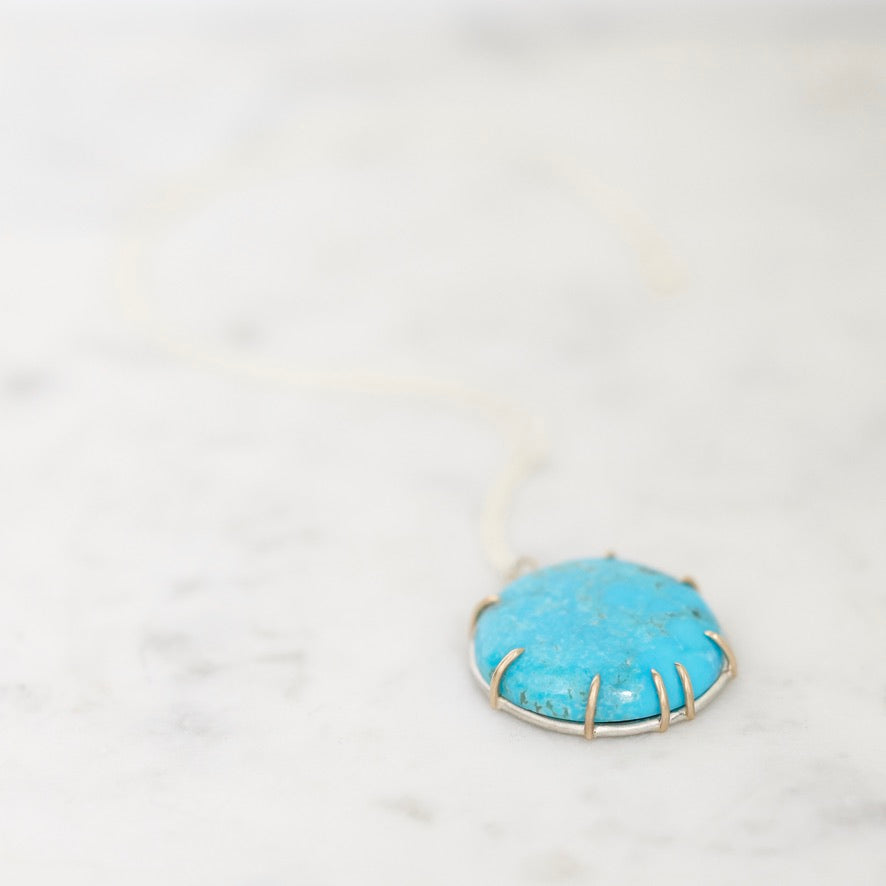 A turquoise stone pendant necklace in a gold-toned bezel setting on a white cord, resting on a light marble surface.