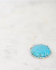 A turquoise stone pendant necklace in a gold-toned bezel setting on a white cord, resting on a light marble surface.