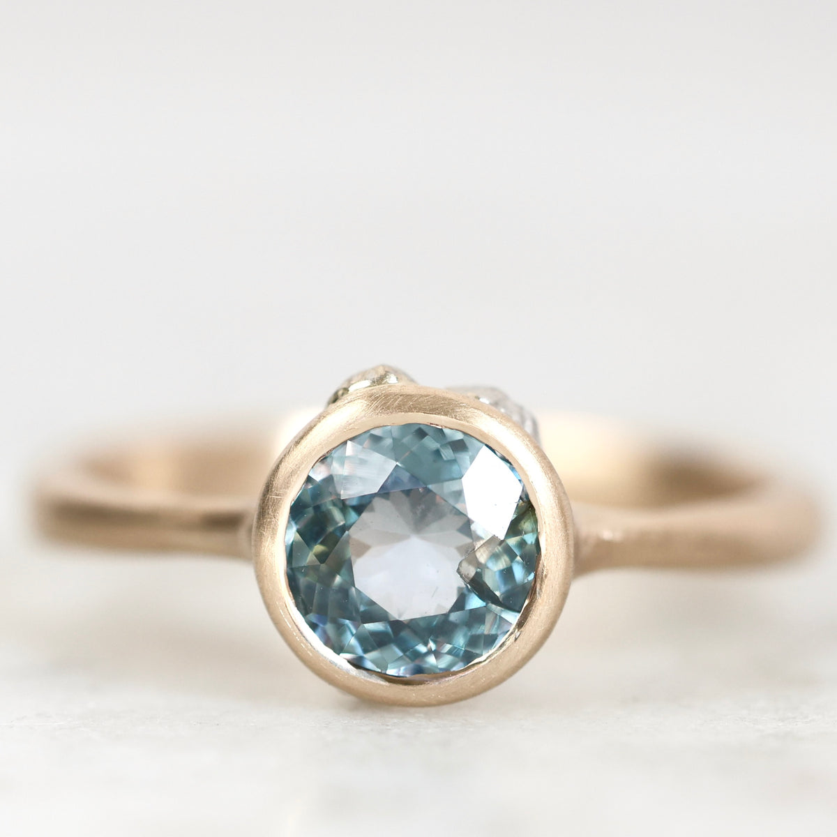A close-up shot of a gold ring featuring a round-cut light blue gemstone in a bezel setting, with a small diamond accent on the side, set against a soft white background.