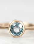 A close-up shot of a gold ring featuring a round-cut light blue gemstone in a bezel setting, with a small diamond accent on the side, set against a soft white background.