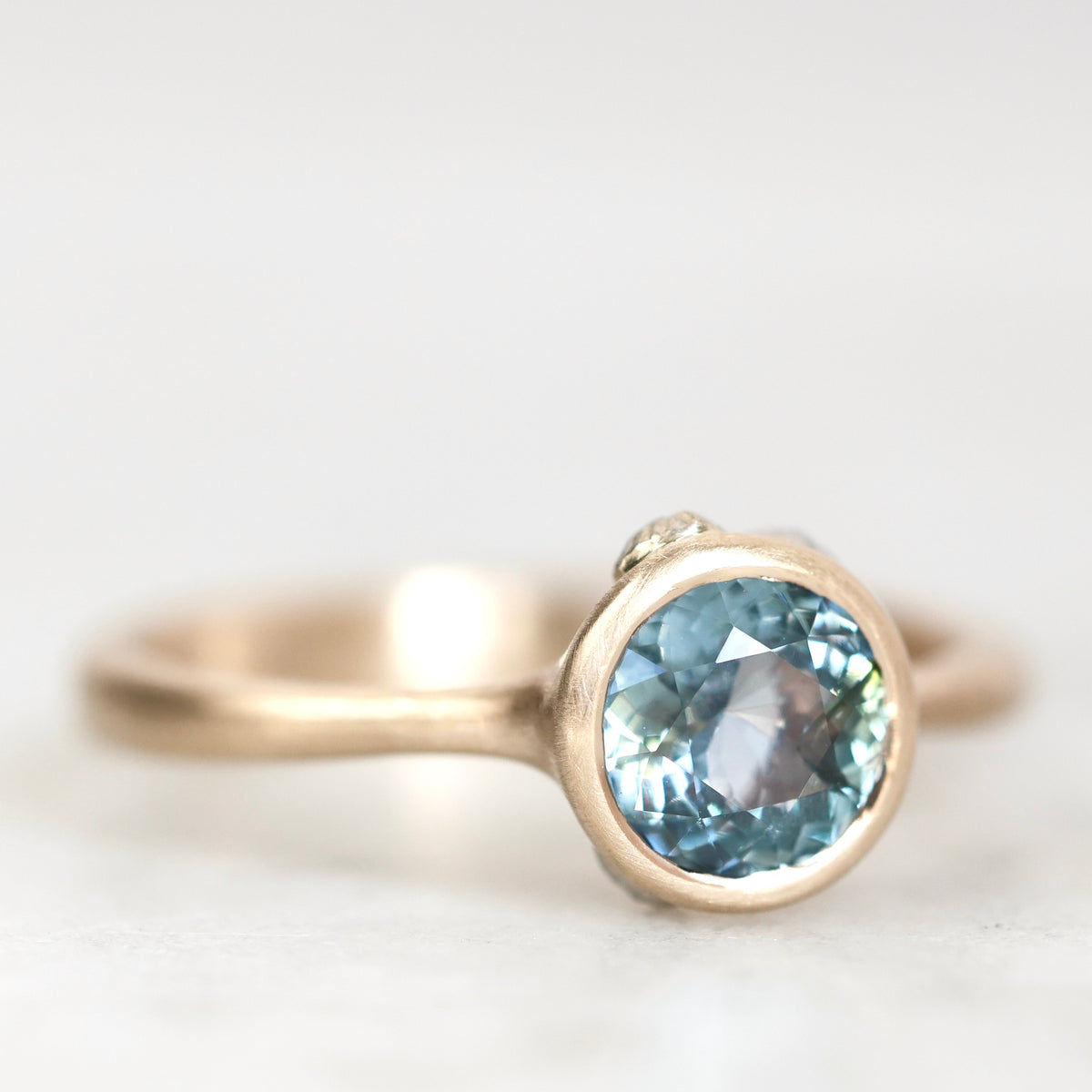 A close-up shot of a gold ring featuring a round-cut light blue gemstone, set against a soft, out-of-focus light gray background.