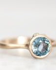 A close-up shot of a gold ring featuring a round-cut light blue gemstone, set against a soft, out-of-focus light gray background.