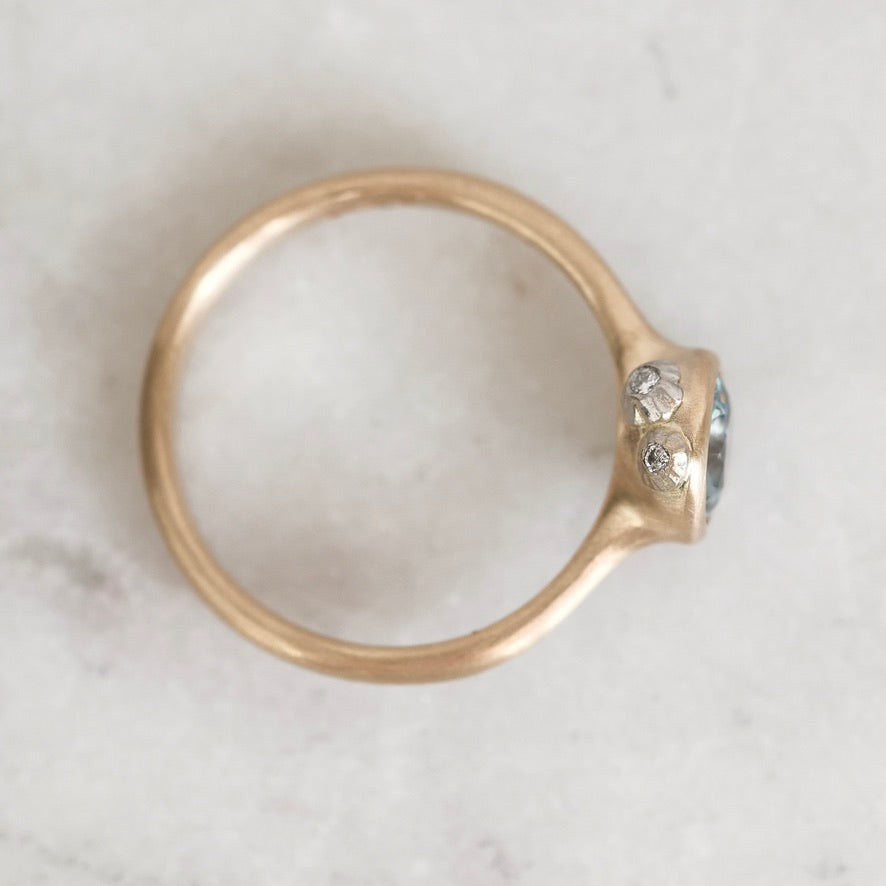 A gold ring with a small cluster of clear gemstones and a tiny blue accent, photographed from above on a light marble surface.