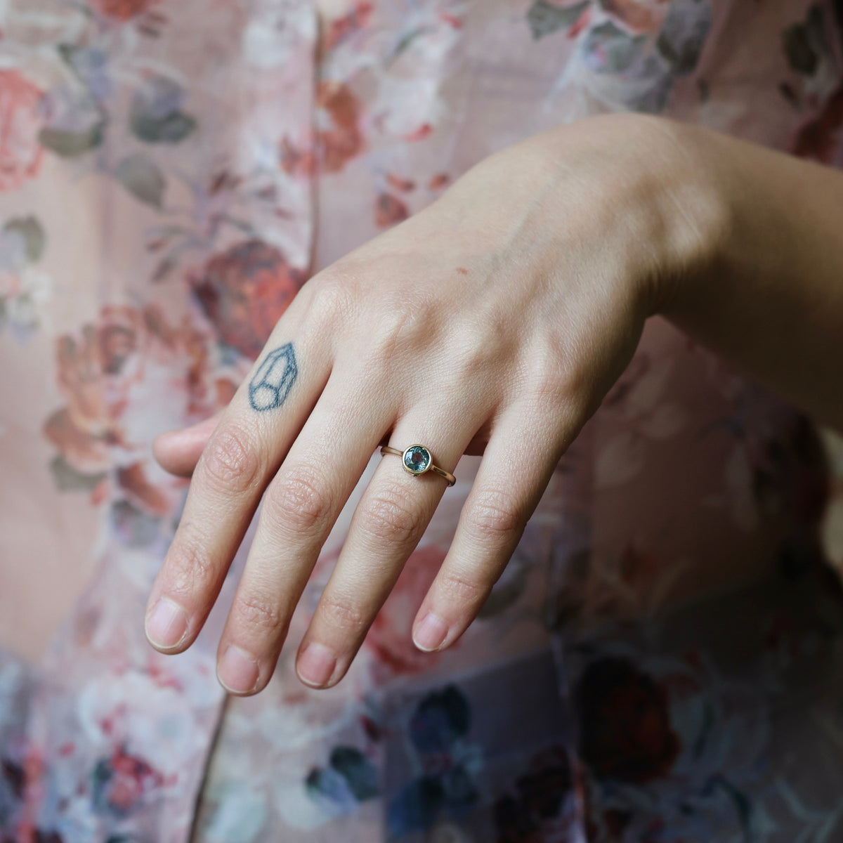 A close-up of a person's hand wearing a delicate gold ring with a small round dark gemstone, set against a soft-focus floral pink background.