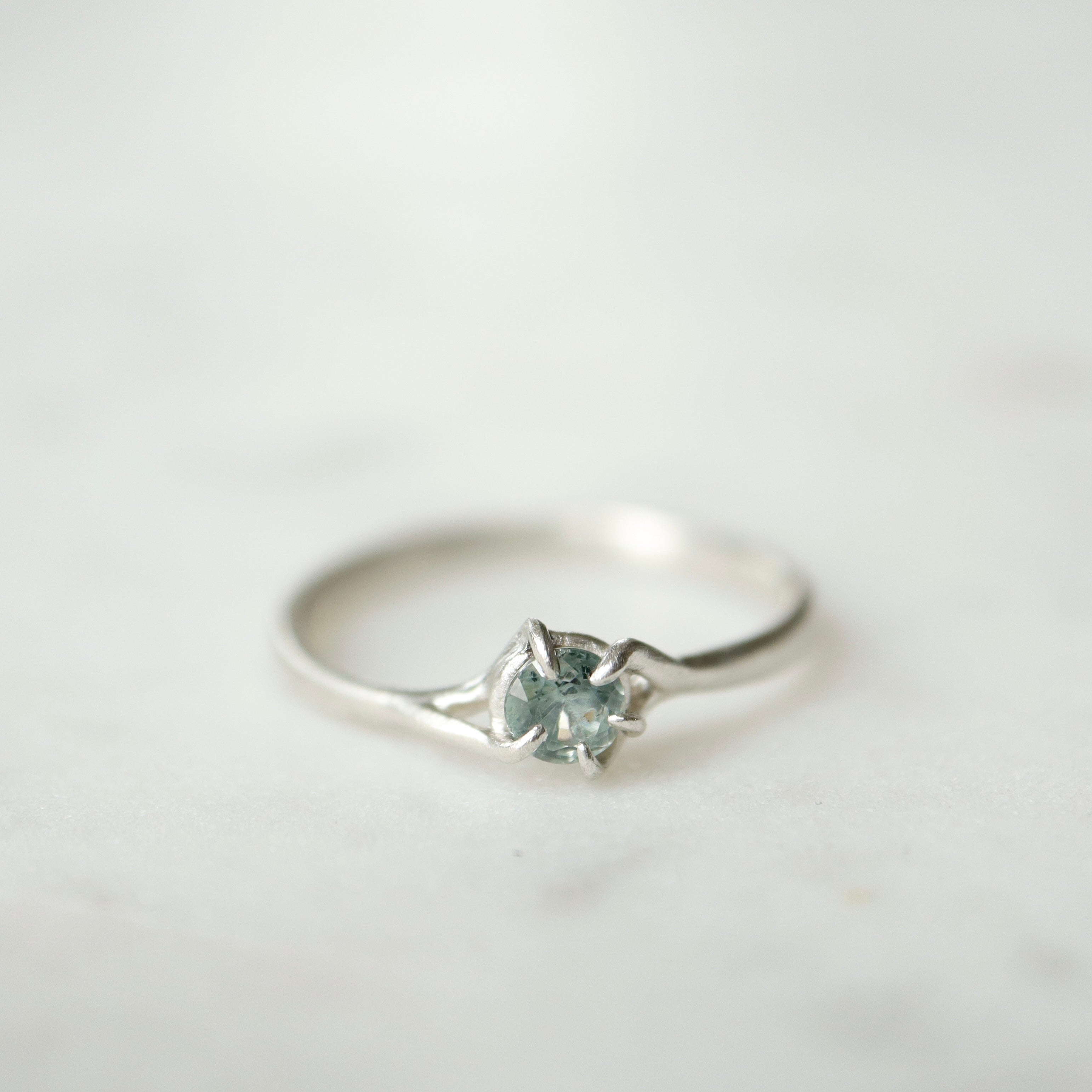 front view of a round pale blue montana sapphire set in a silver ring. The stone is wrapped in branching prongs. Shown on a white marble slab.