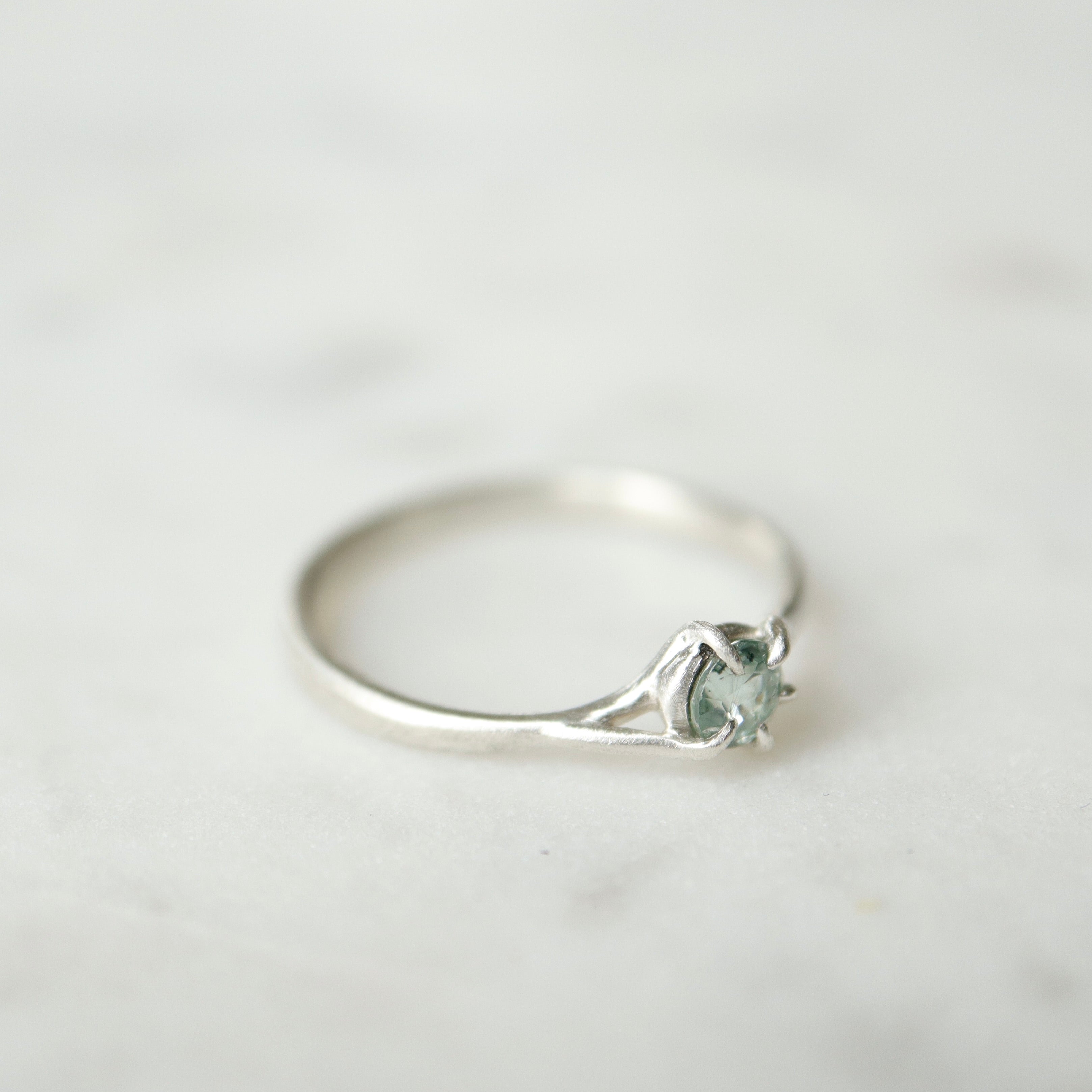 side view of a round pale blue montana sapphire set in a silver ring. The stone is wrapped in branching prongs. Shown on a white marble slab.