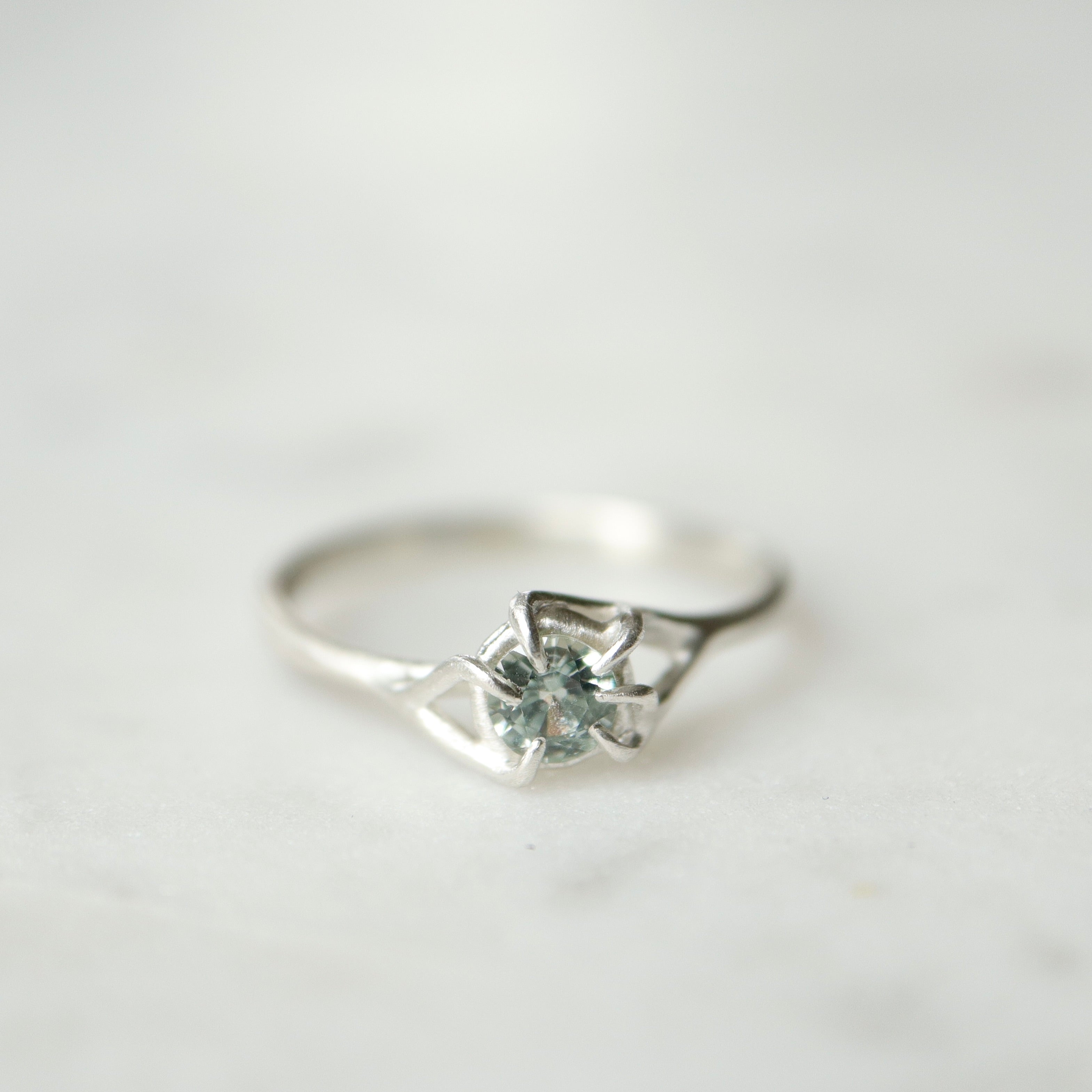 Front view of a round, pale blue montana sapphire set in a silver ring. The stone is wrapped in branching prongs and photographed on a white marble slab.