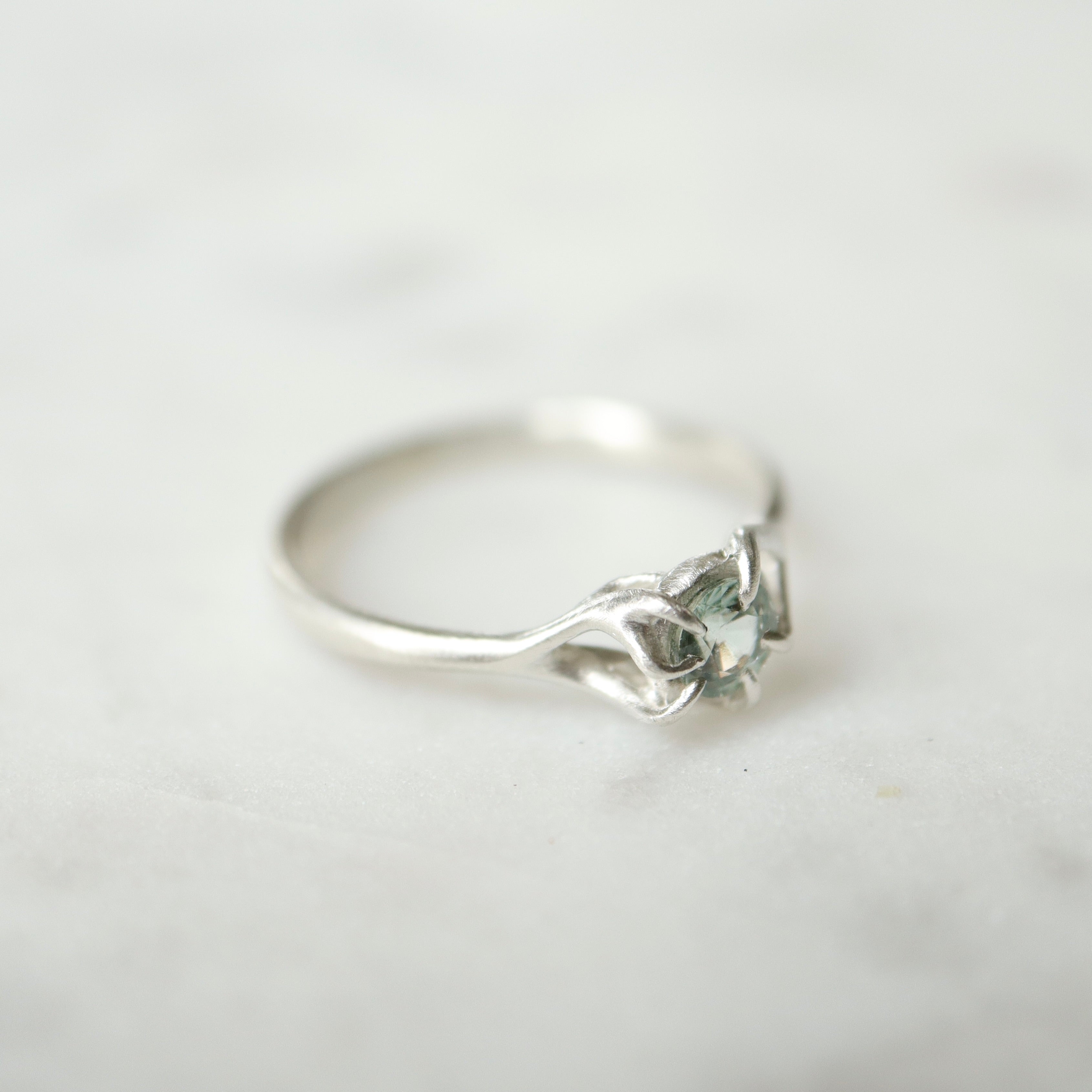Side view of a round, pale blue montana sapphire set in a silver ring. The stone is wrapped in branching prongs and photographed on a white marble slab.