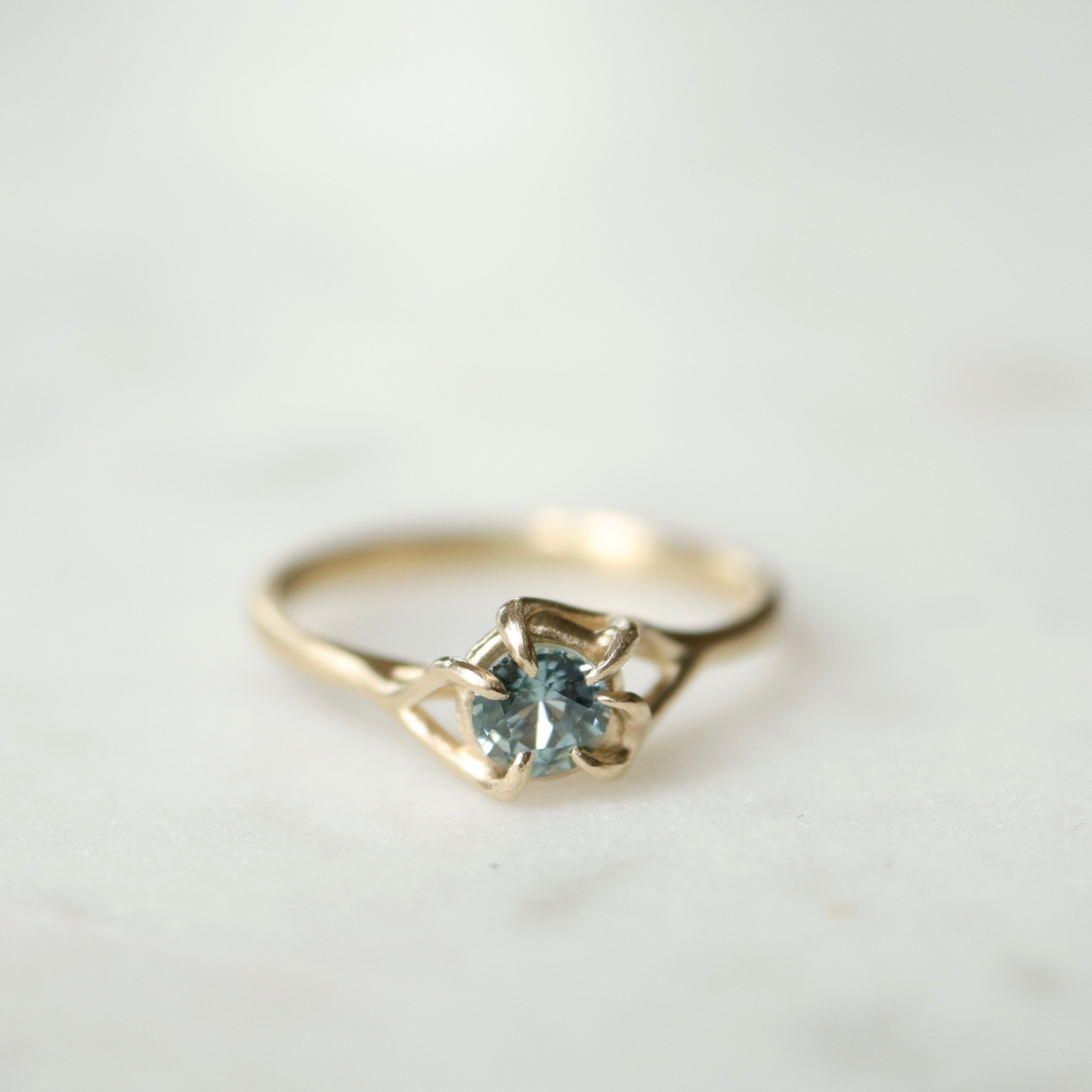 front view of a gold ring. a round crystal blue montana sapphire is wrapped in gold branches. photographed on a white marble slab.