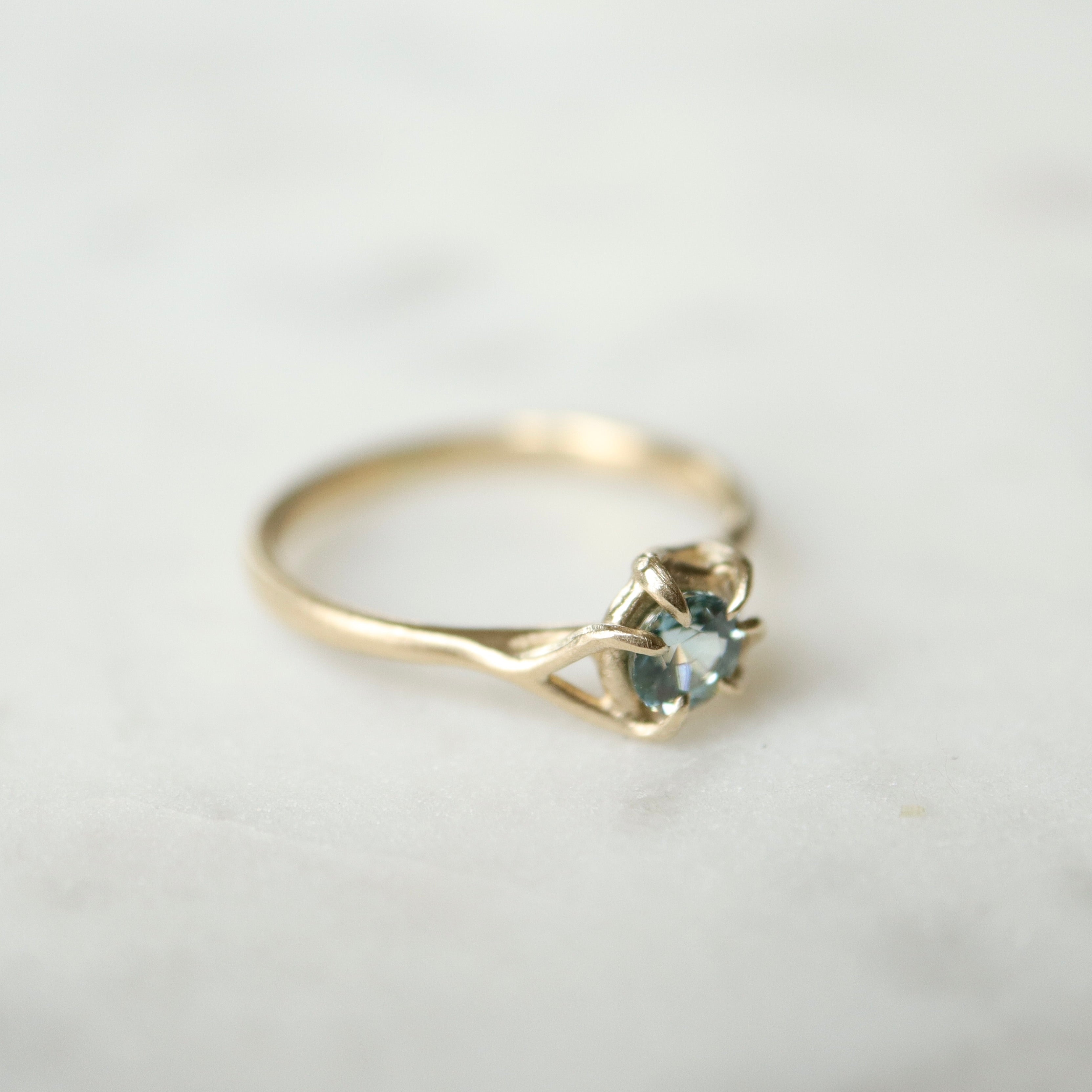 side view of a gold ring. a round crystal blue montana sapphire is wrapped in gold branches. photographed on a white marble slab.