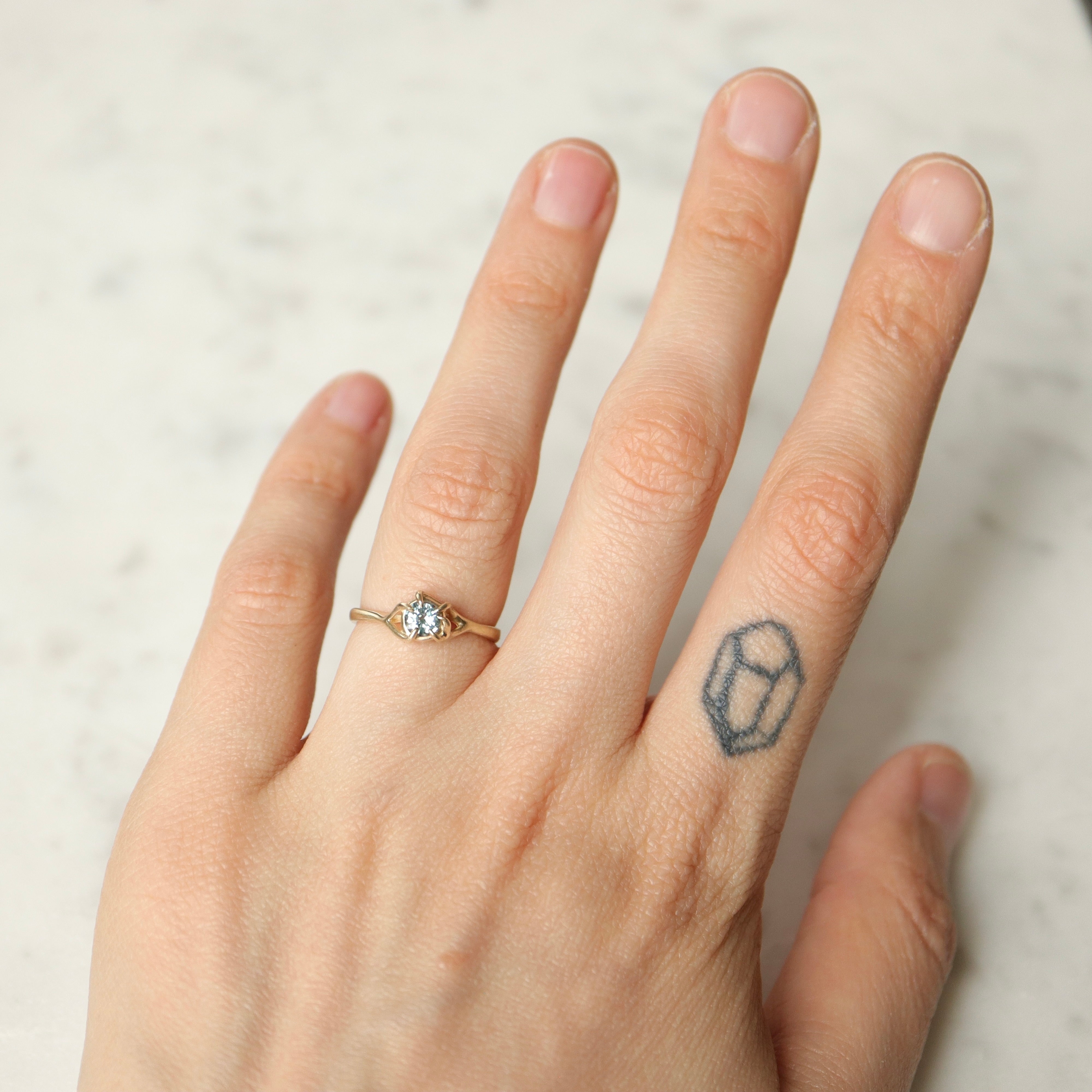 on-hand view of a gold ring. a round crystal blue montana sapphire is wrapped in gold branches. photographed on a white marble slab.