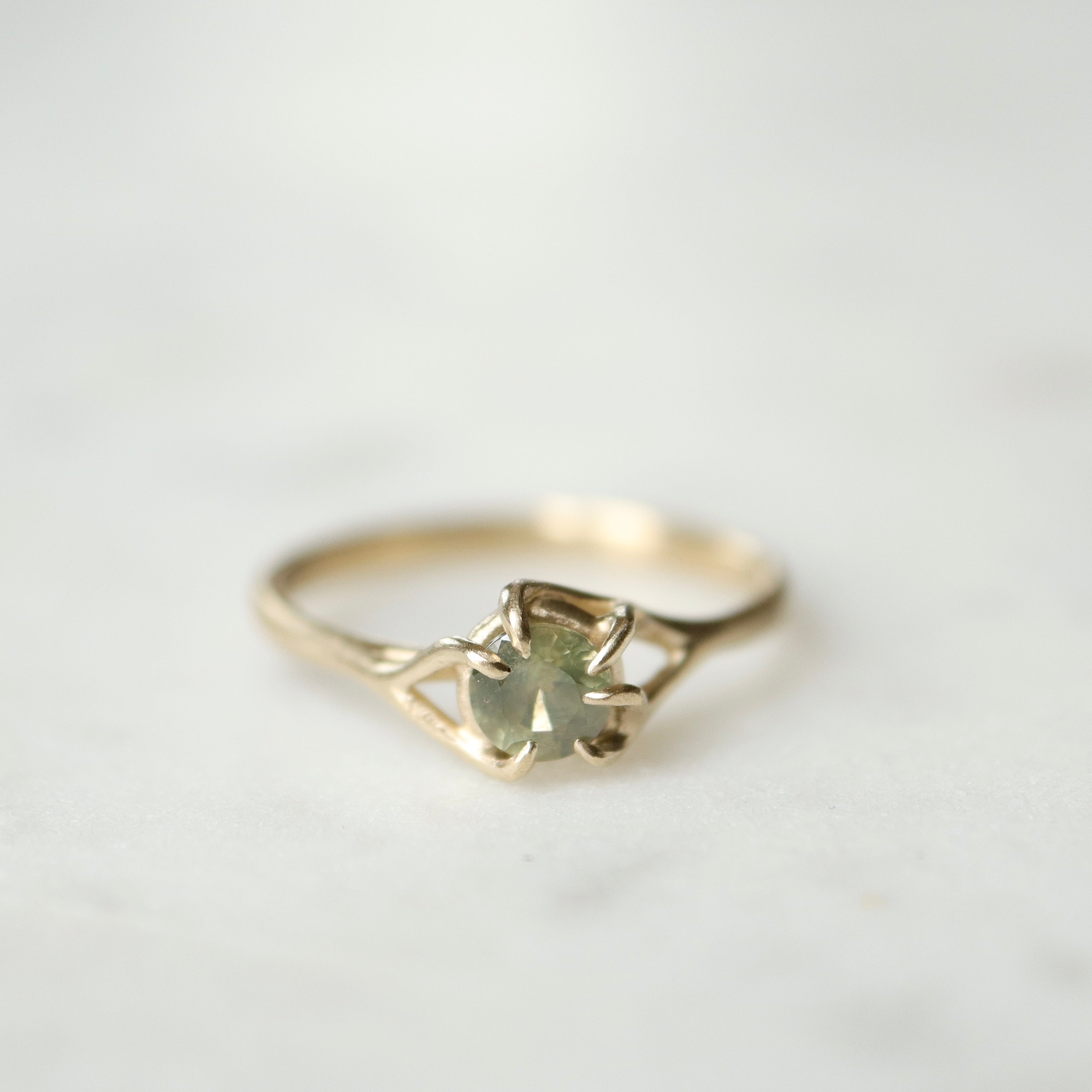 front view of a round sage green montana sapphire set in a gold ring with branching prongs. Shown on a white marble surface.