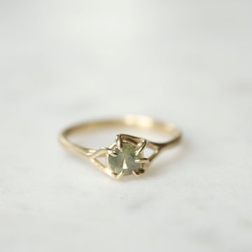 front view of a round sage green montana sapphire set in a gold ring with branching prongs. Shown on a white marble surface.