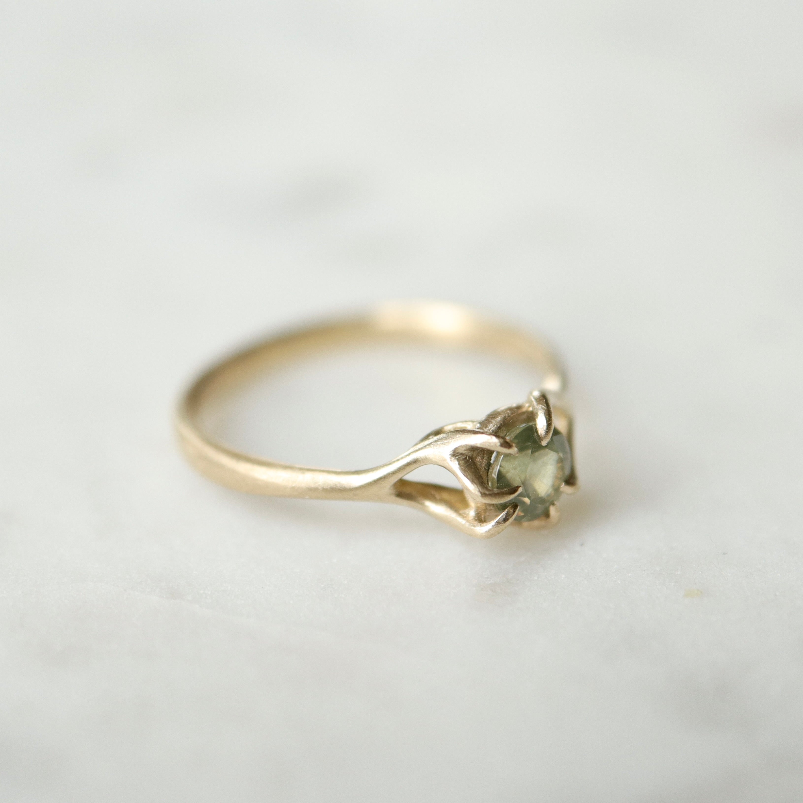 side view of a round sage green montana sapphire set in a gold ring with branching prongs. Shown on a white marble surface.