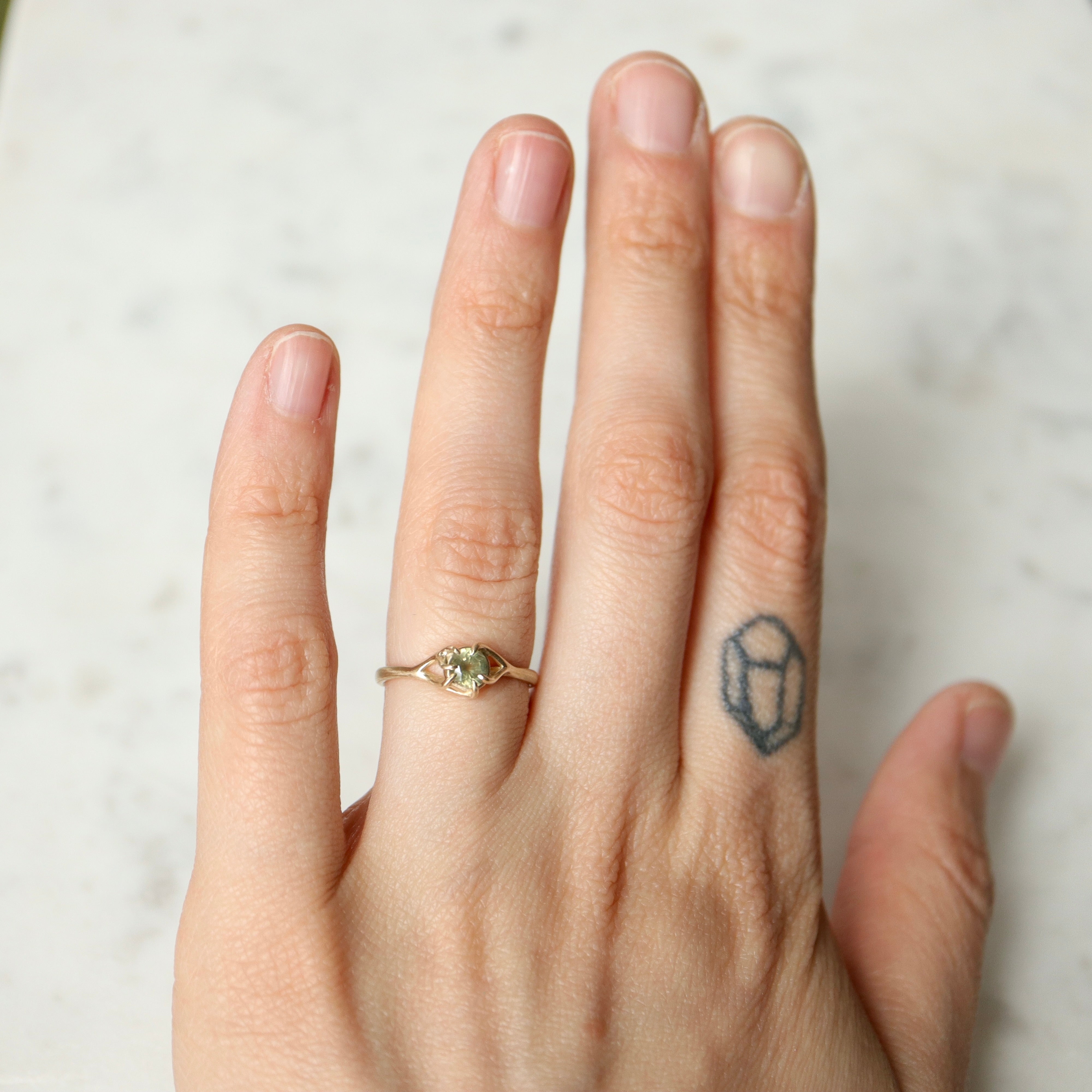 on-hand view of a round sage green montana sapphire set in a gold ring with branching prongs. Shown on a white marble surface.