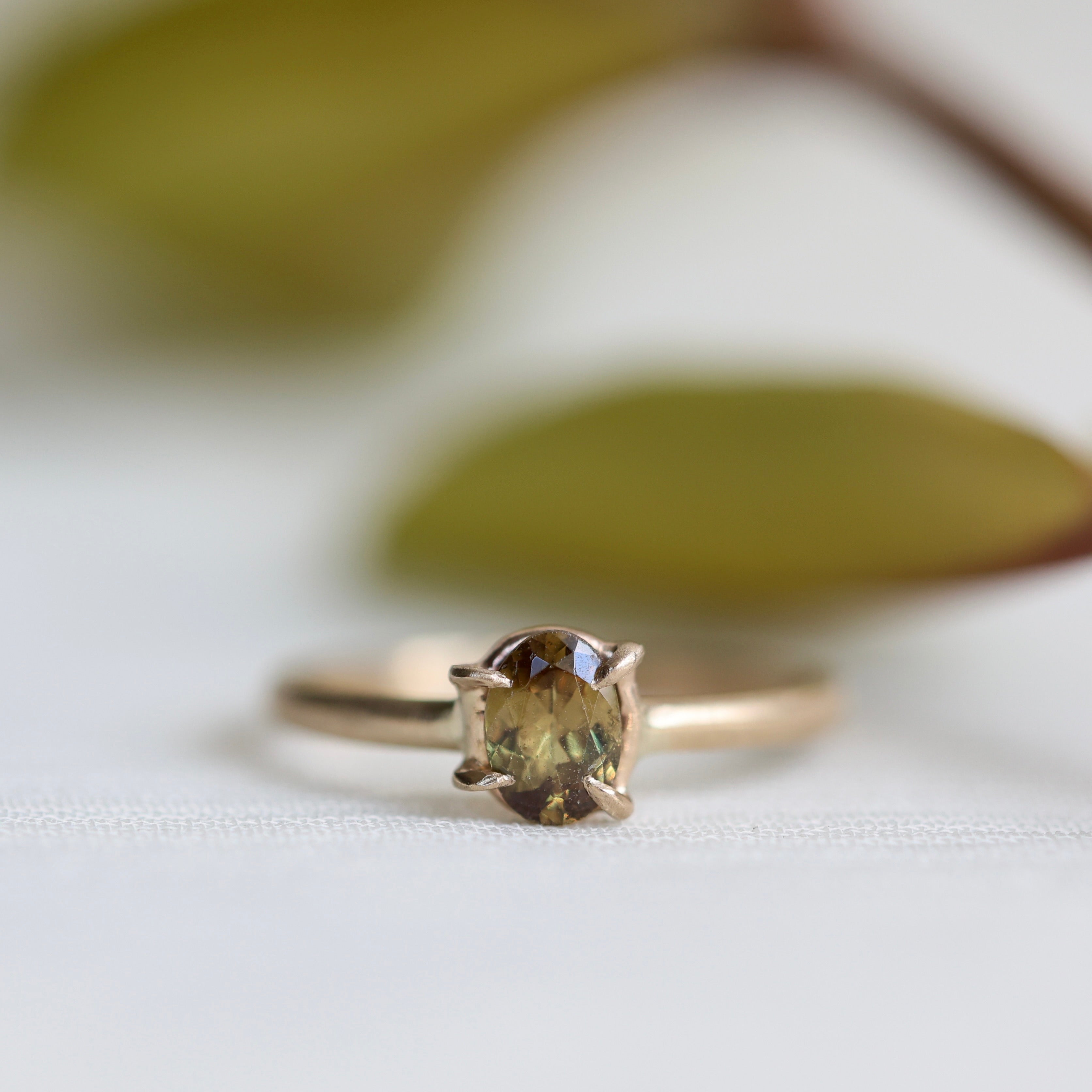 Gold ring with a brownish green gemstone on a white background with faded leaves