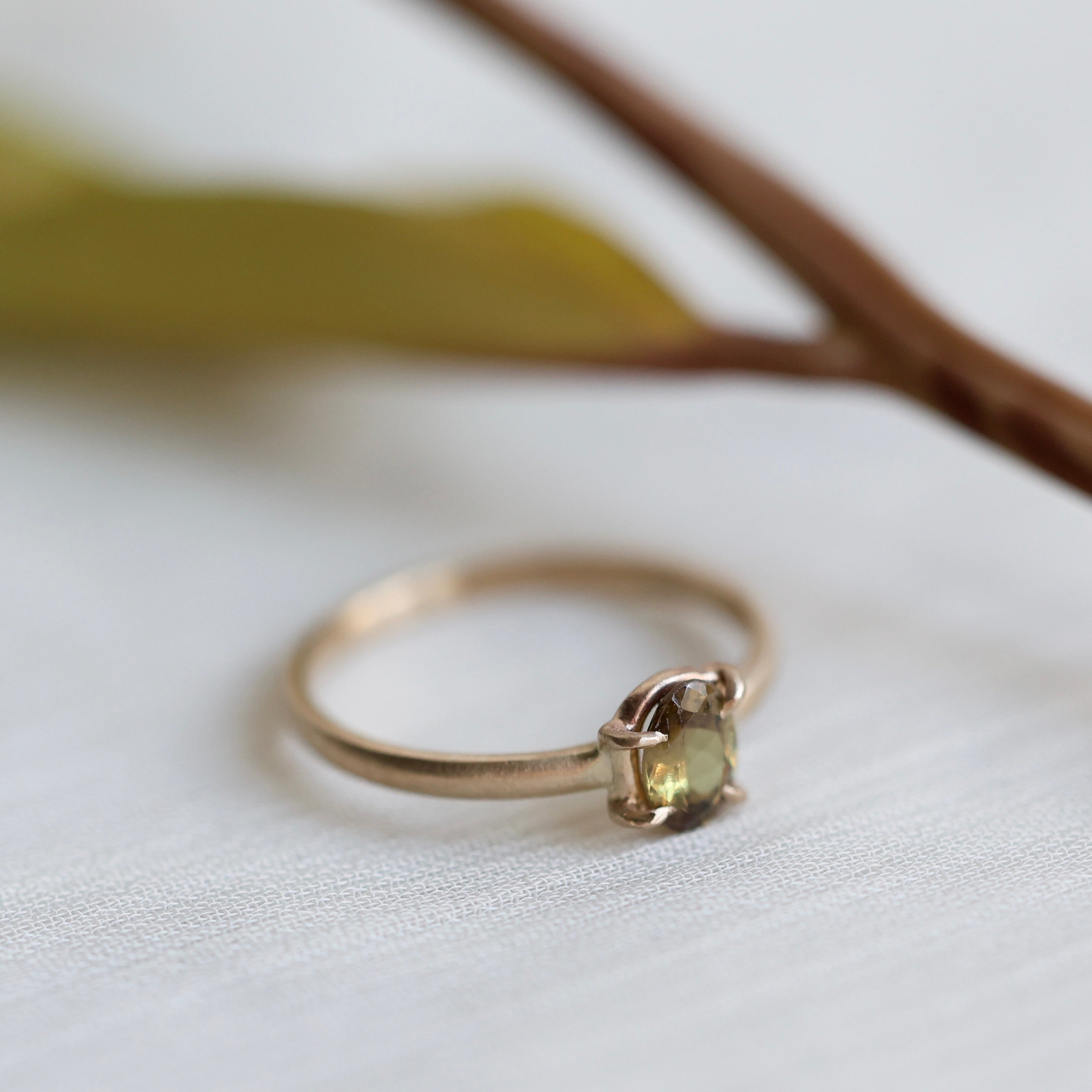 Gold ring with a brownish green gemstone on a white background with faded leaves