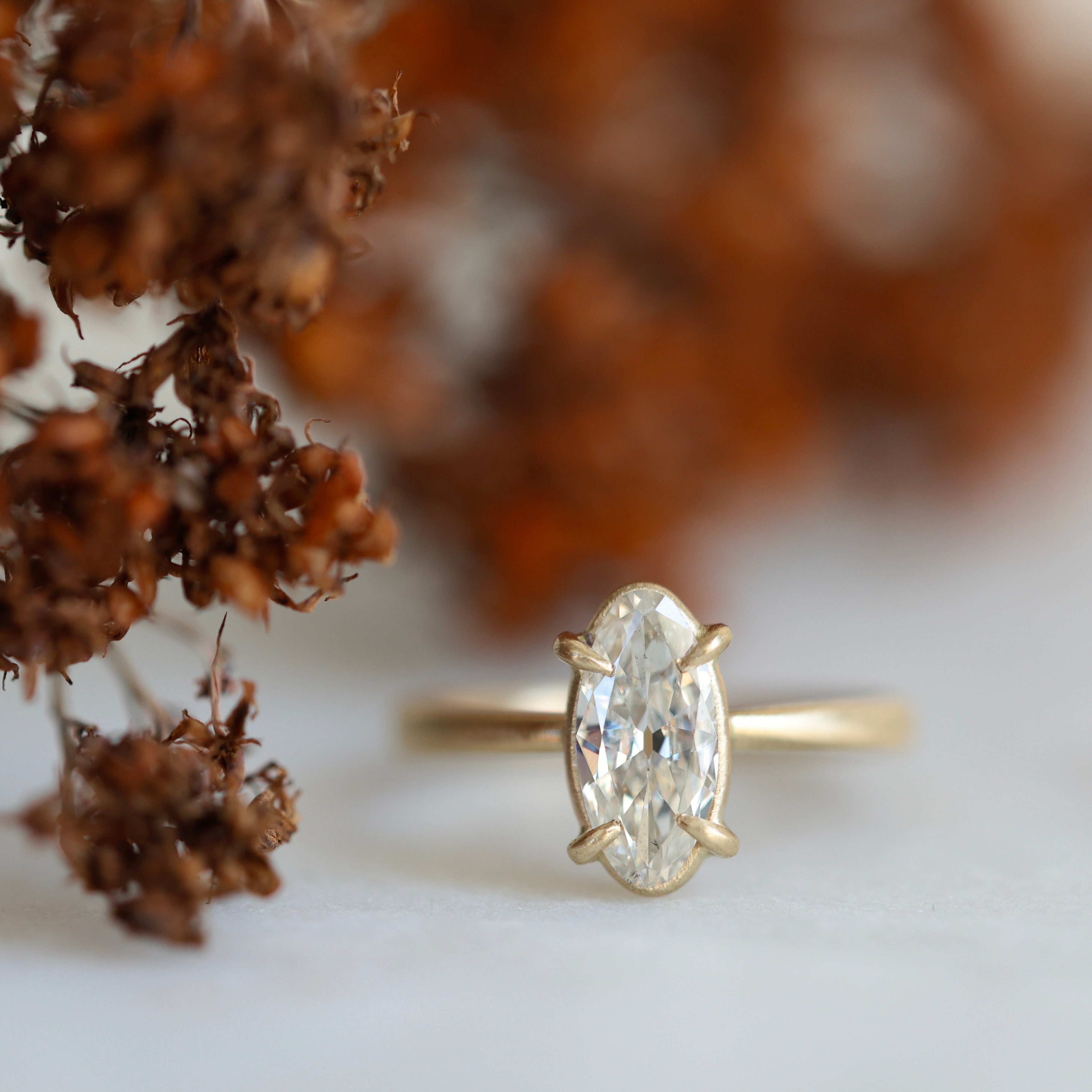 elongated oval diamond set in yellow gold ring with prongs. photographed with brown flowers