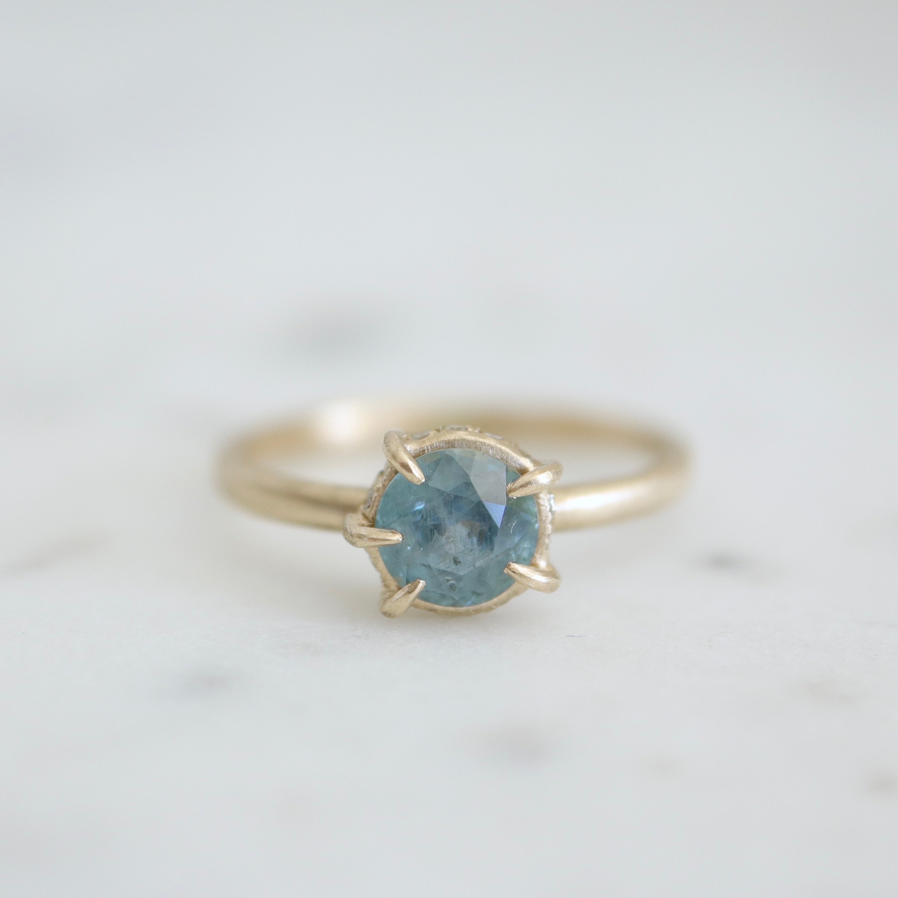 a muted teal round sapphire sits within gold prongs in this gold ring, a hidden halo of diamonds beneath. Front view.