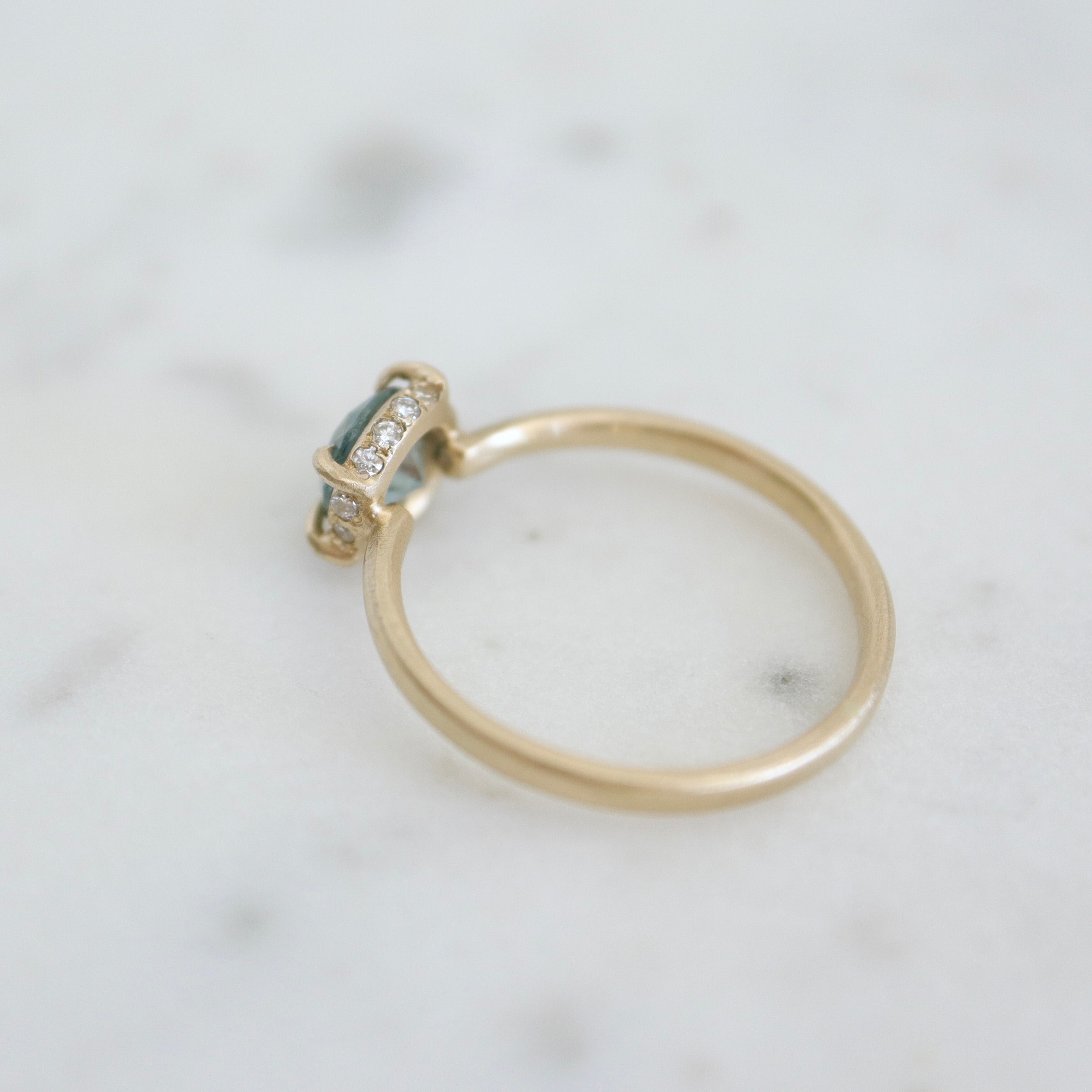 a muted teal round sapphire sits within gold prongs in this gold ring, a hidden halo of diamonds beneath. back view.