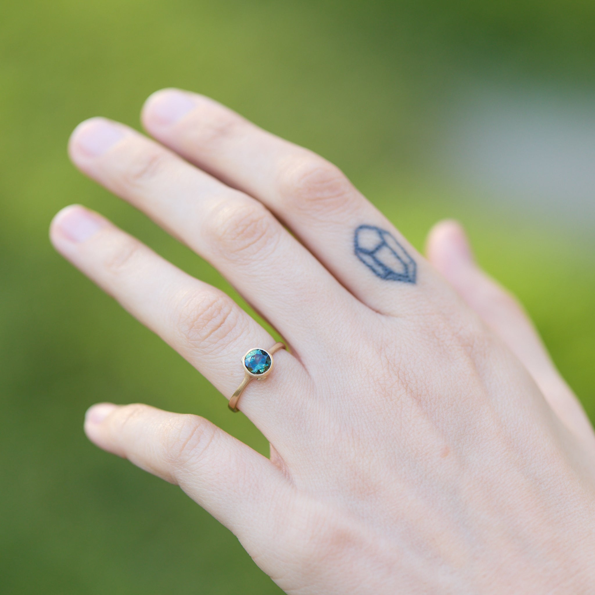 Hand wearing a ring with a blue gemstone against a blurred green background