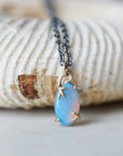 closeup of oval opal pendant with blue, green and pink flashes, set in gold branch prongs, on oxidized silver black chain. Set against a fossil shell background. necklace By hannah blount