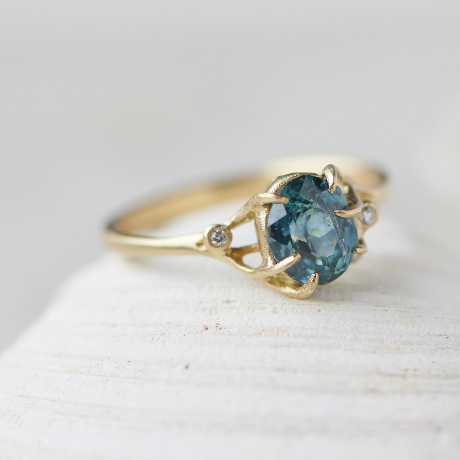 oval teal montana sapphire is set in a gold branch ring with vine-like prongs holding the stone in. Three little diamond berries adorn the branches. 1/4 view.