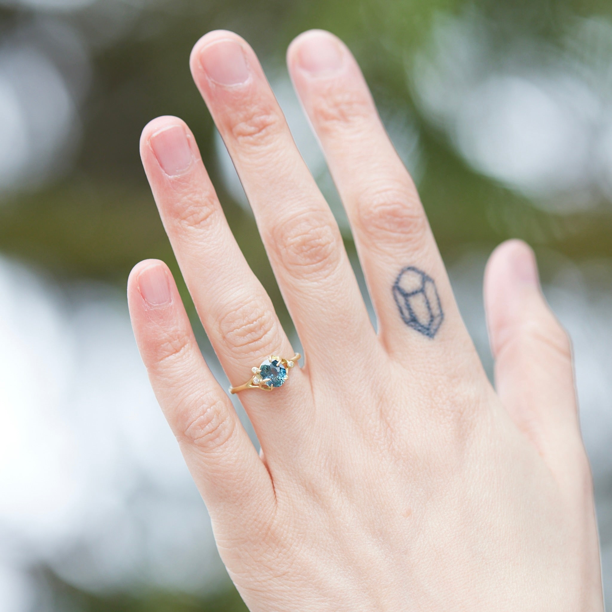 oval teal montana sapphire is set in a gold branch ring with vine-like prongs holding the stone in. Three little diamond berries adorn the branches. Photographed on hand.