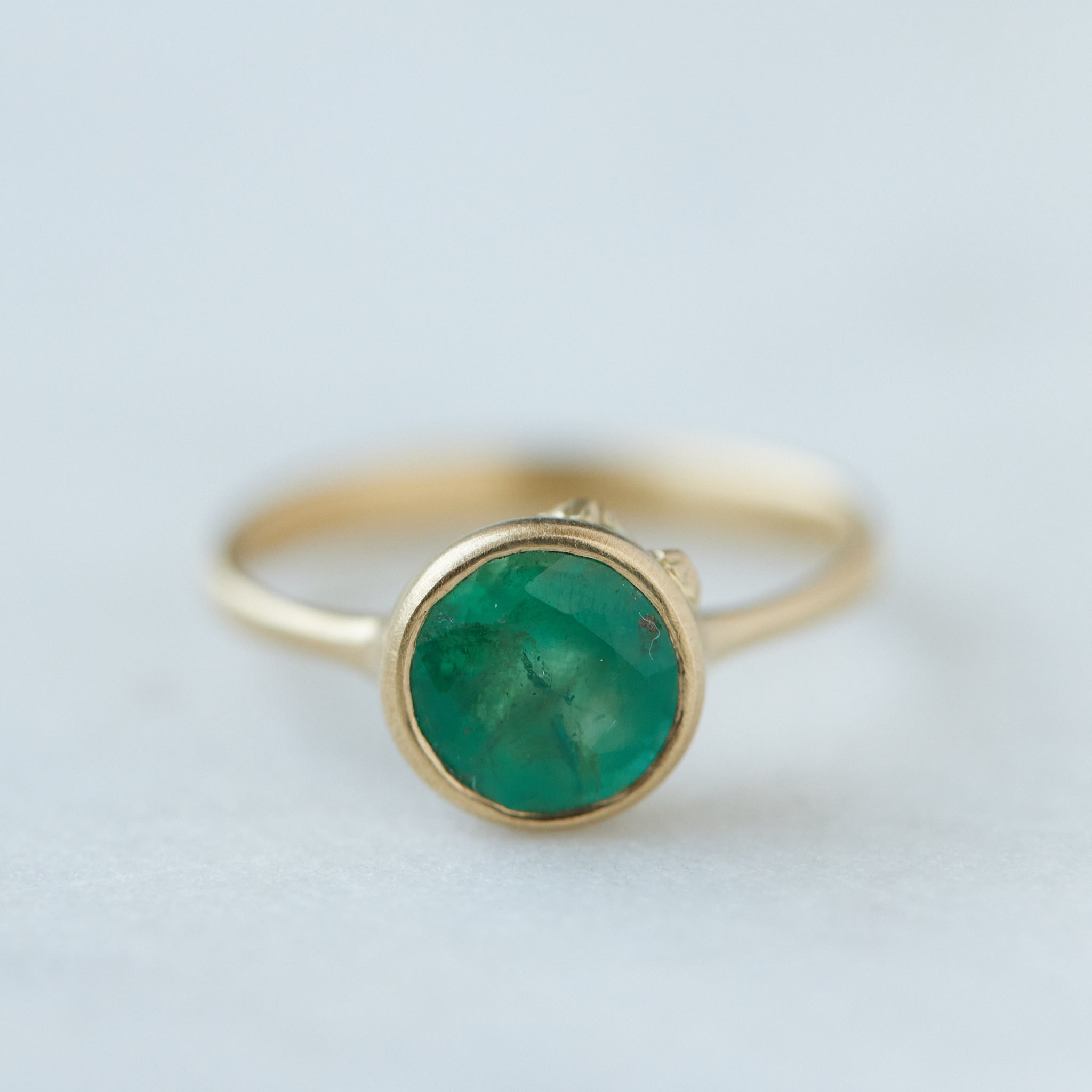 round rich green emerald is set in a gold bezel ring with gold barnacles. Front view.