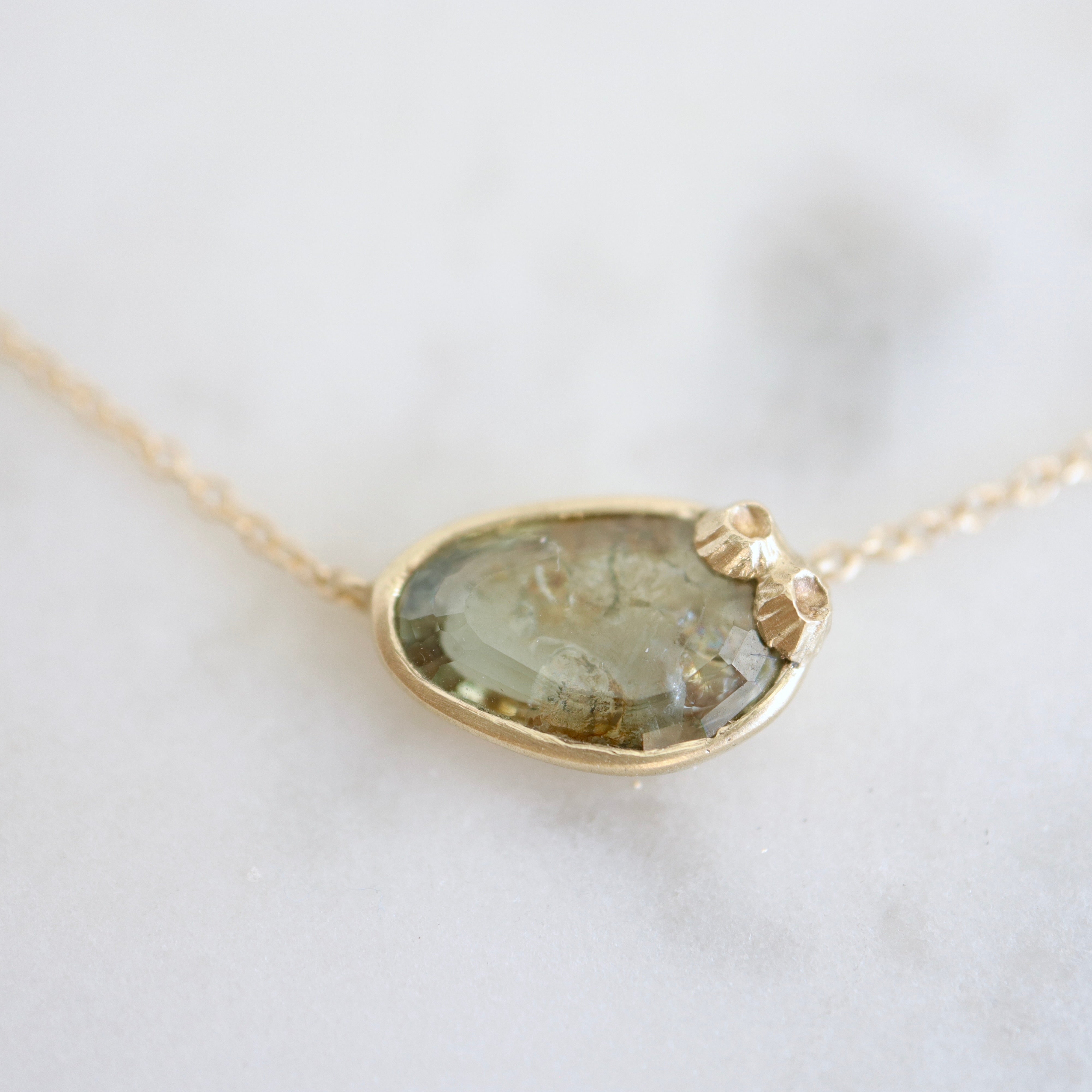 a green and brown a-symmetrical sapphire is set in a gold bezel on a gold chain, with two little barnacles in the right corner. close-up view.