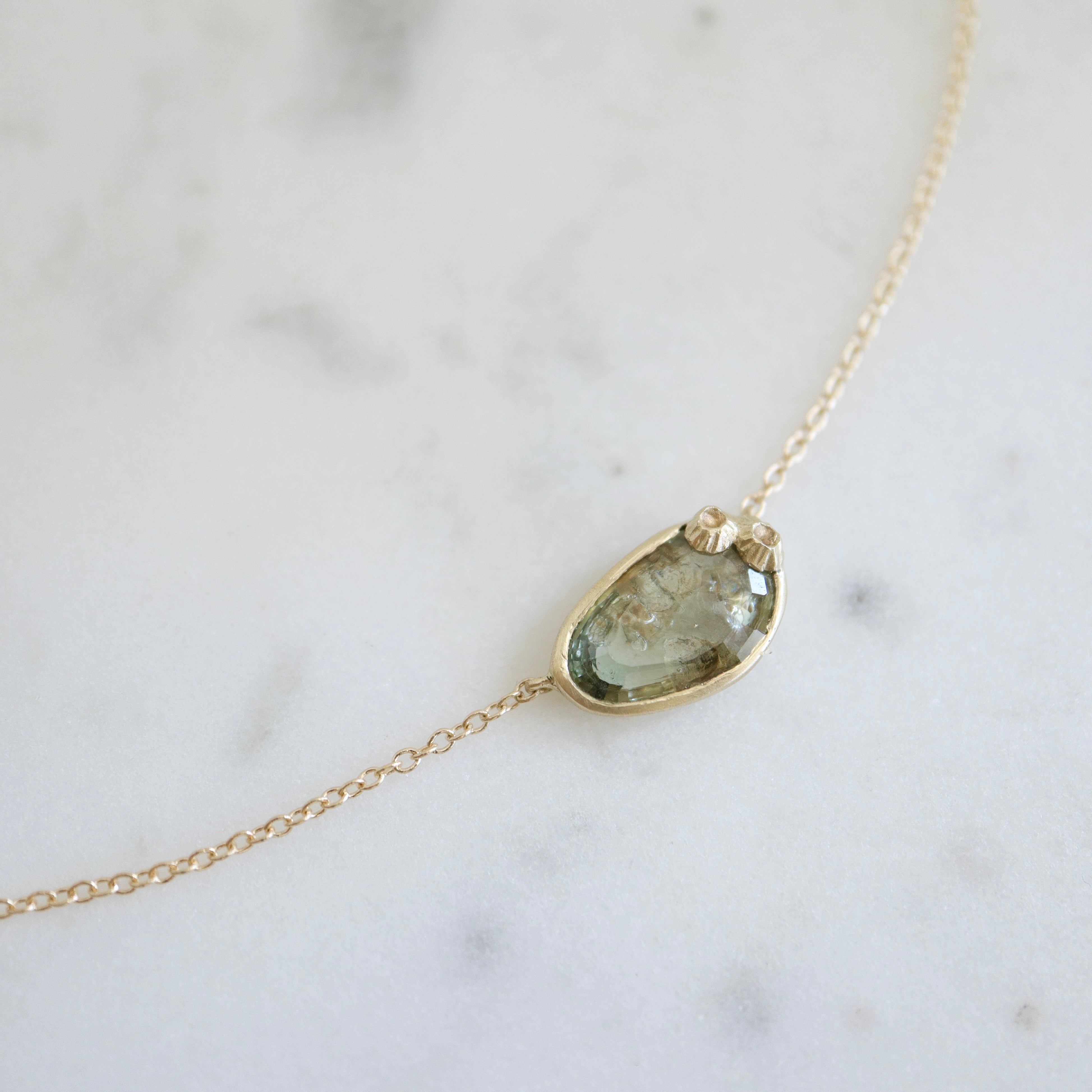 a green and brown a-symmetrical sapphire is set in a gold bezel on a gold chain, with two little barnacles in the right corner. detail view.