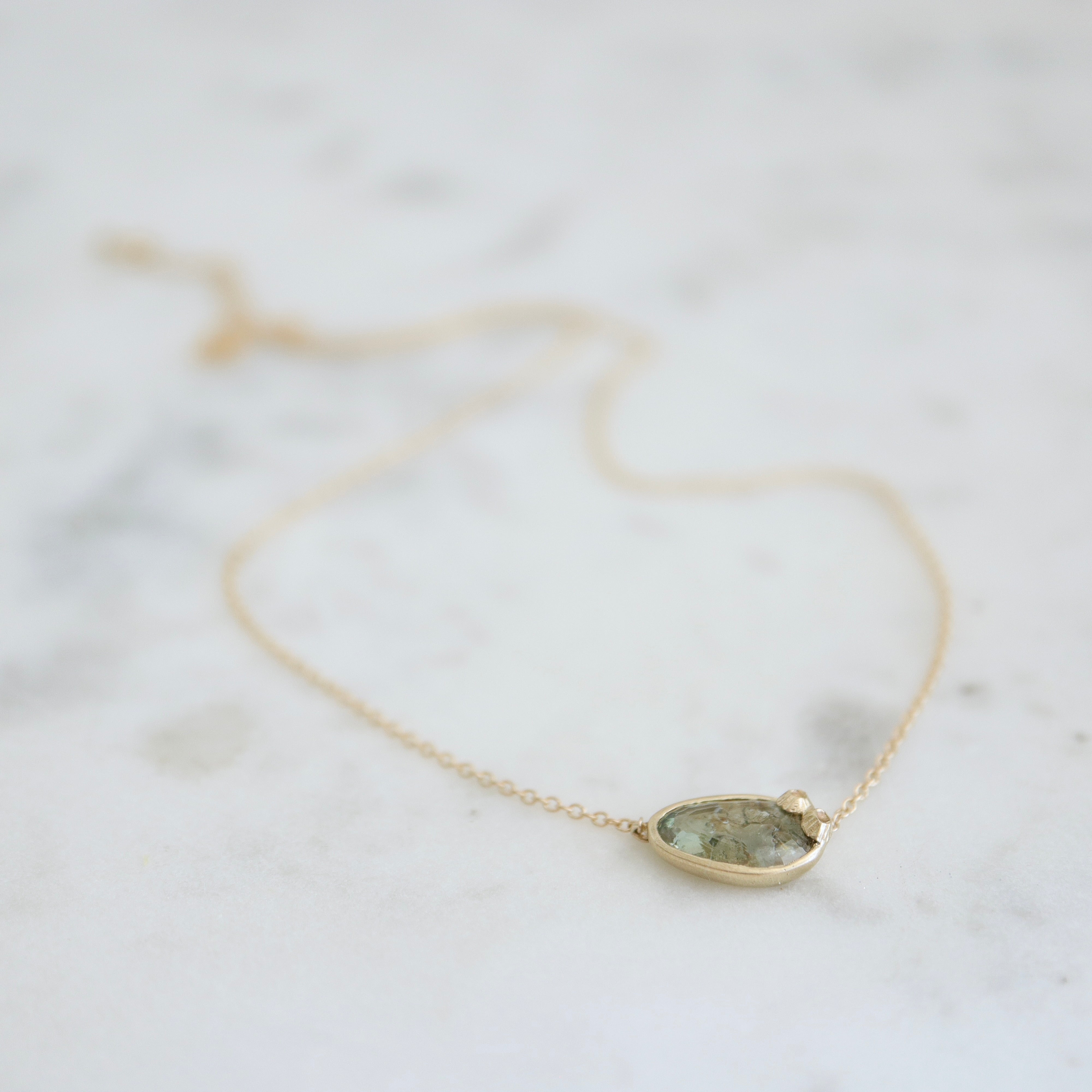 a green and brown a-symmetrical sapphire is set in a gold bezel on a gold chain, with two little barnacles in the right corner. whole view.