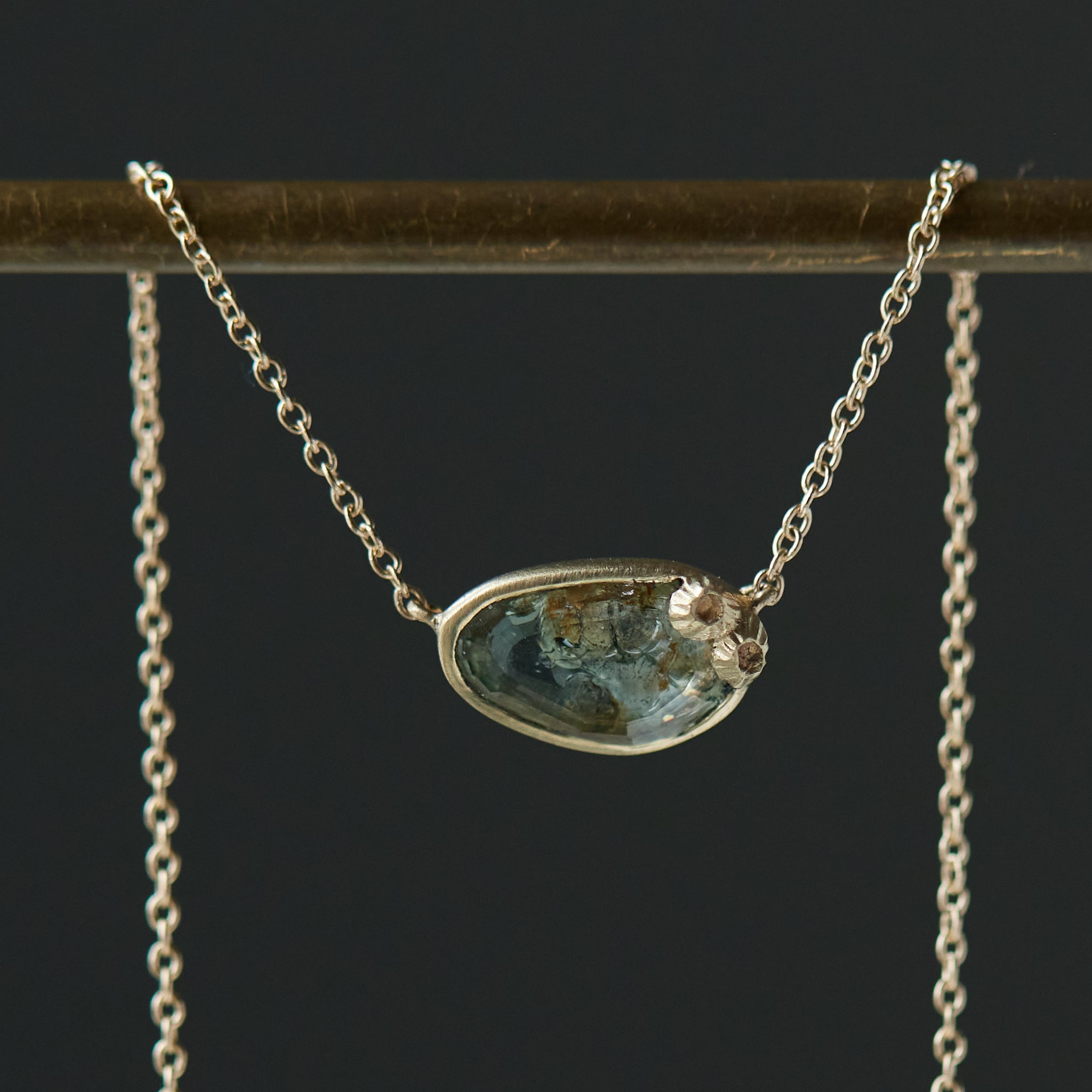 a green and brown a-symmetrical sapphire is set in a gold bezel on a gold chain, with two little barnacles in the right corner. close-up view.