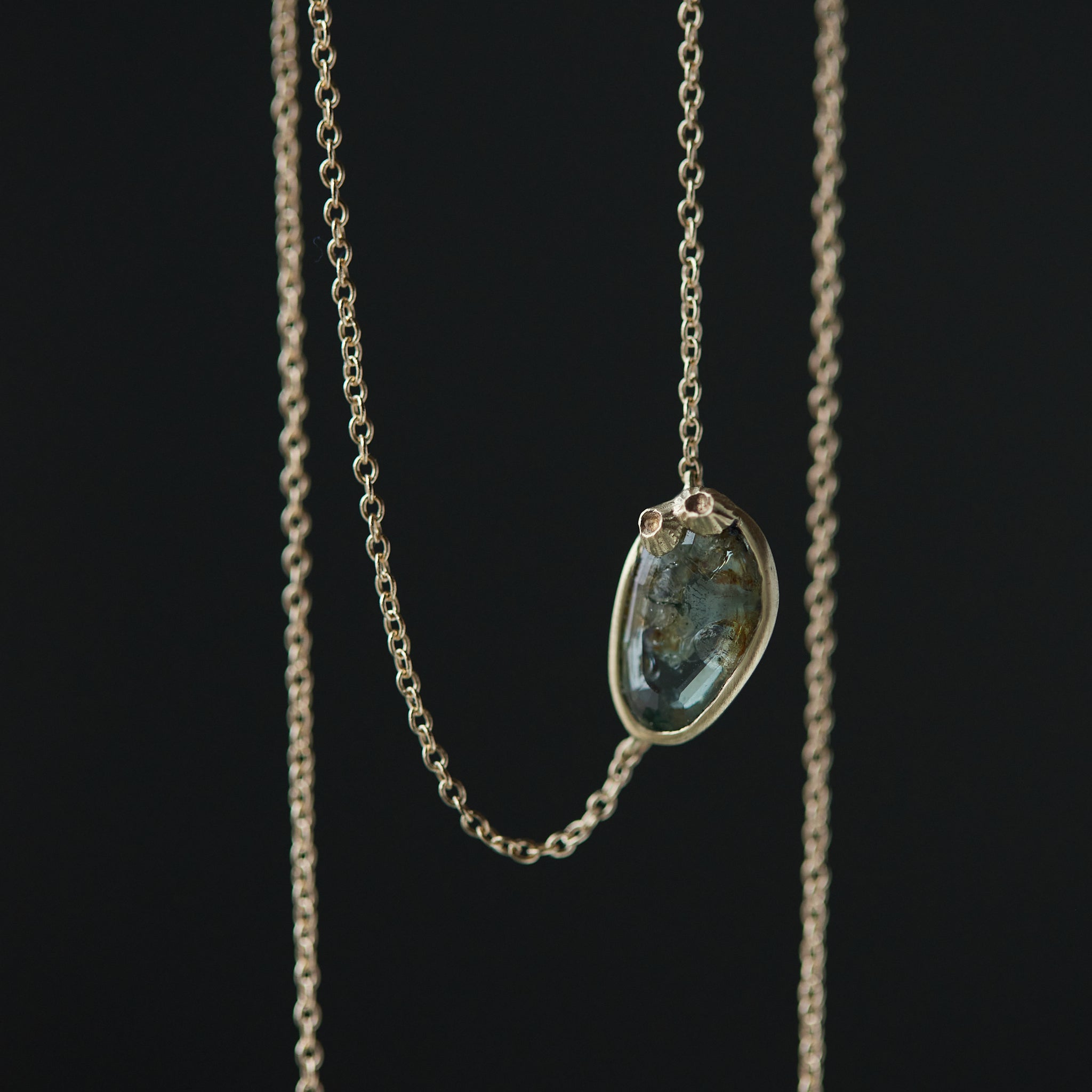 a green and brown a-symmetrical sapphire is set in a gold bezel on a gold chain, with two little barnacles in the right corner. close-up view.