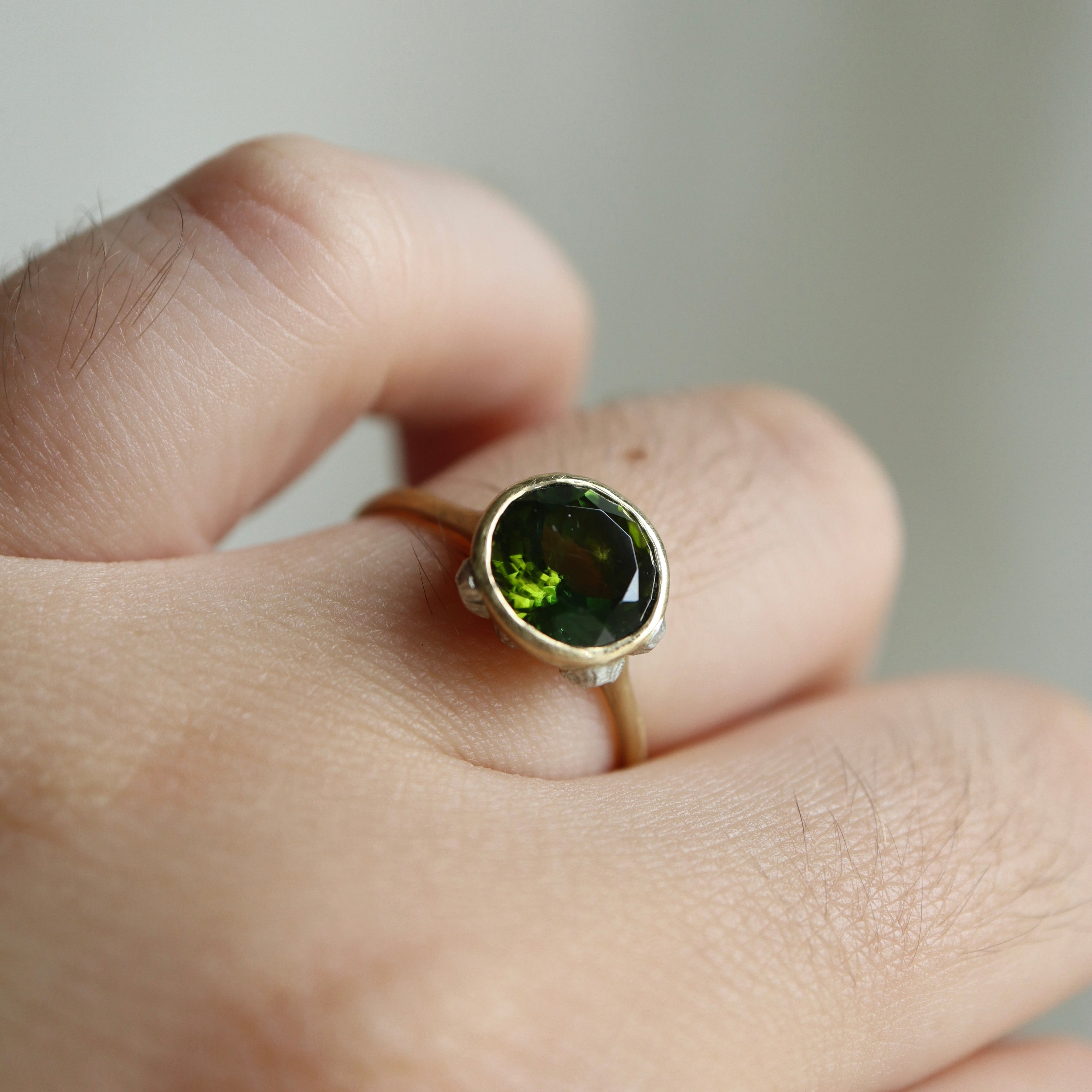 14k yellow gold bezel set ring with a green Tourmaline and bright sterling silver barnacles on a finger against a neutral background.