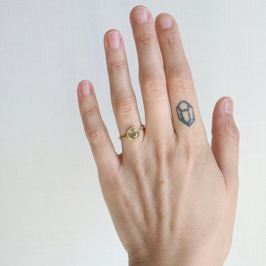 Hand wearing a gold ring with a small diamond-shaped tattoo on the finger against a plain background