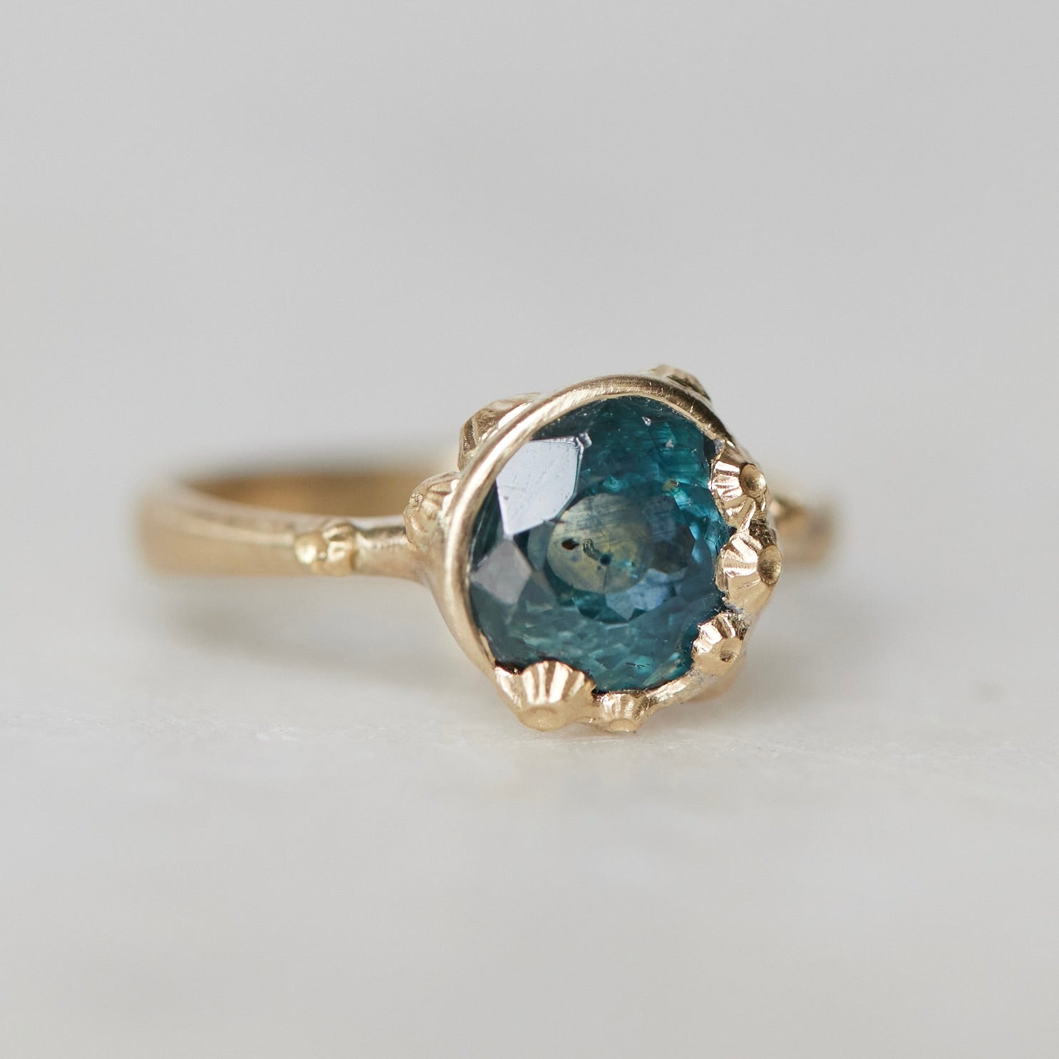 a deep teal montana sapphire is set in a gold bezel of a gold ring, with barnacles coming up over the side of the bezel and onto the stone, as well as along the band. 1/4 view.