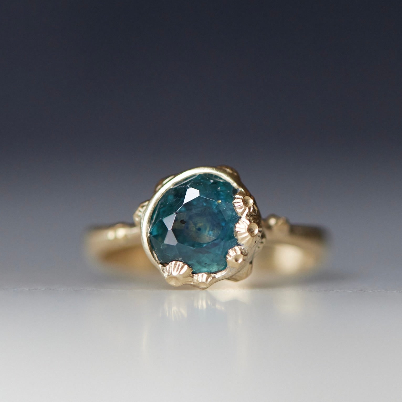 a deep teal montana sapphire is set in a gold bezel of a gold ring, with barnacles coming up over the side of the bezel and onto the stone, as well as along the band. front view.