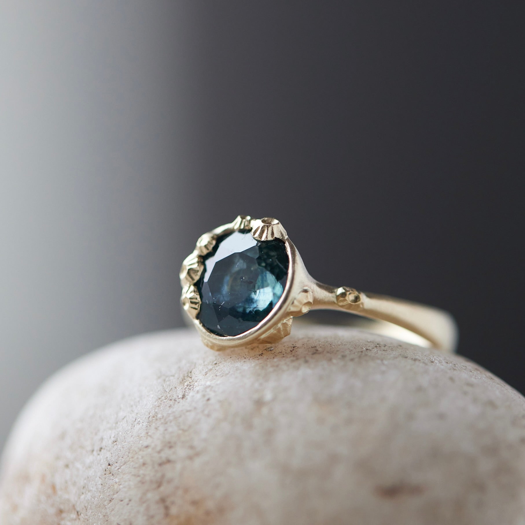 a deep teal montana sapphire is set in a gold bezel of a gold ring, with barnacles coming up over the side of the bezel and onto the stone, as well as along the band. 1/4 view.
