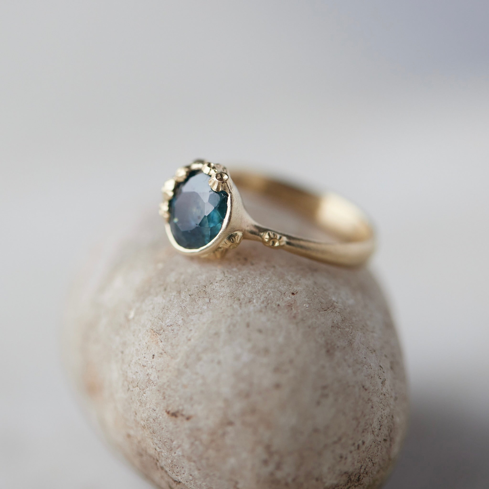 a deep teal montana sapphire is set in a gold bezel of a gold ring, with barnacles coming up over the side of the bezel and onto the stone, as well as along the band. side view.