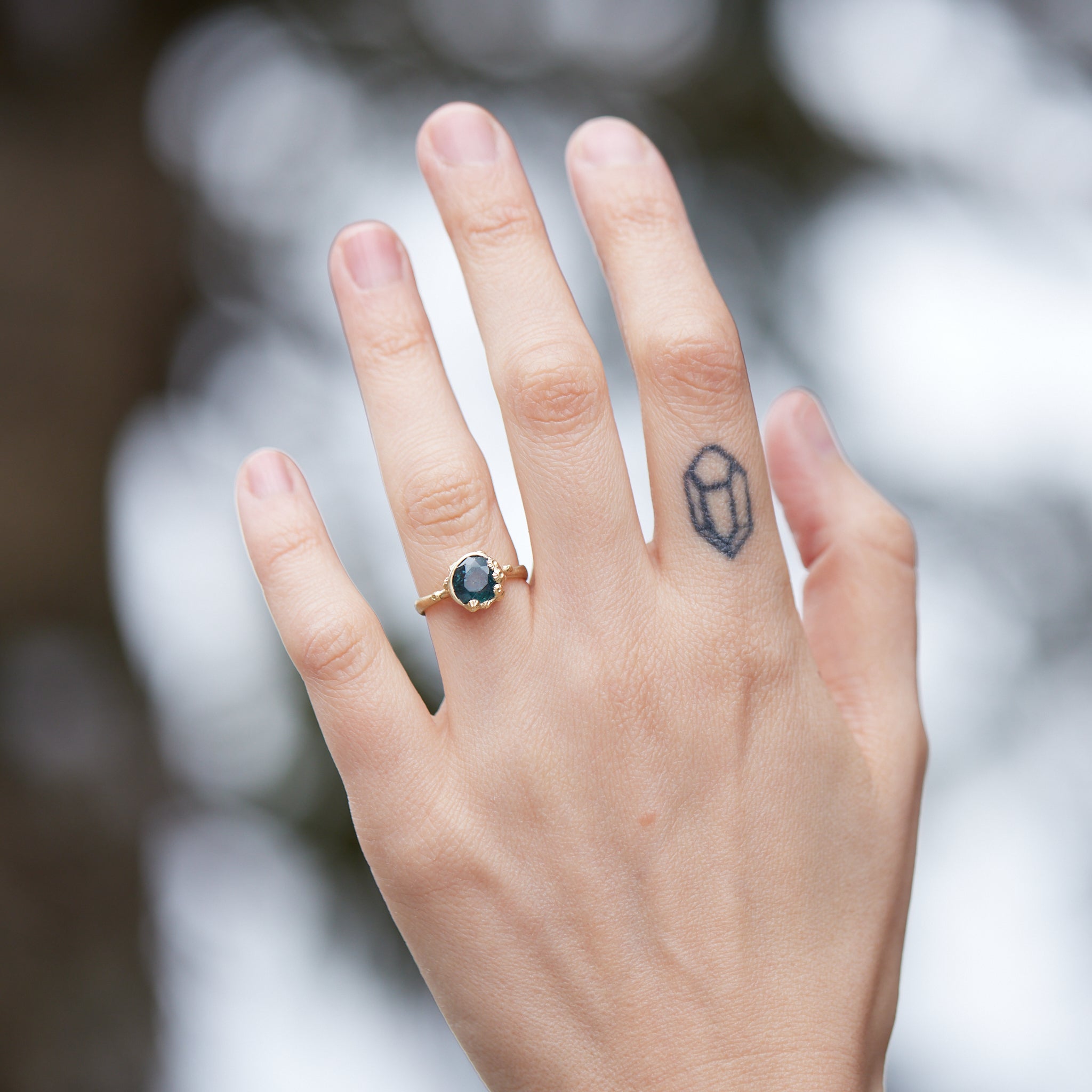 a deep teal montana sapphire is set in a gold bezel of a gold ring, with barnacles coming up over the side of the bezel and onto the stone, as well as along the band. photographed on hand.