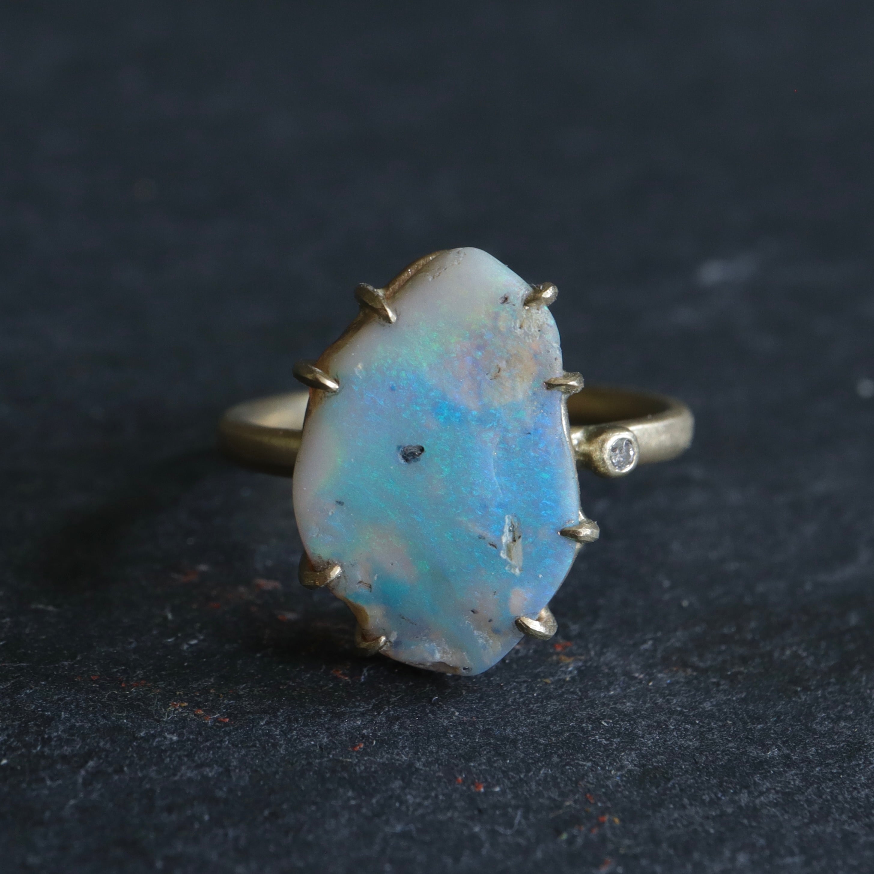 raw opal with rainbow colors is set within eight gold prongs on a gold band. A single diamond sits atop the band next to the opal, like a little friend.