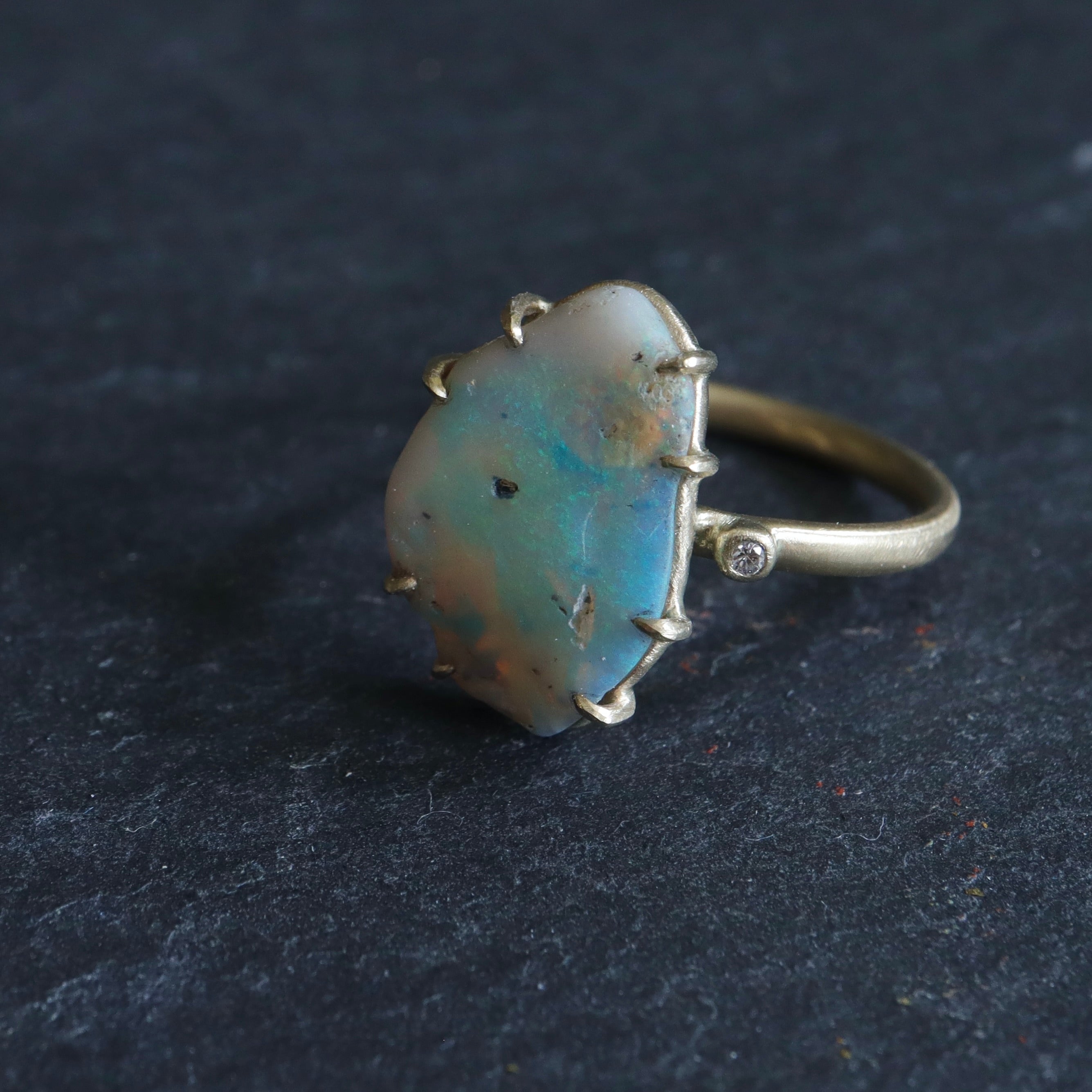 raw opal with rainbow colors is set within eight gold prongs on a gold band. A single diamond sits atop the band next to the opal, like a little friend.